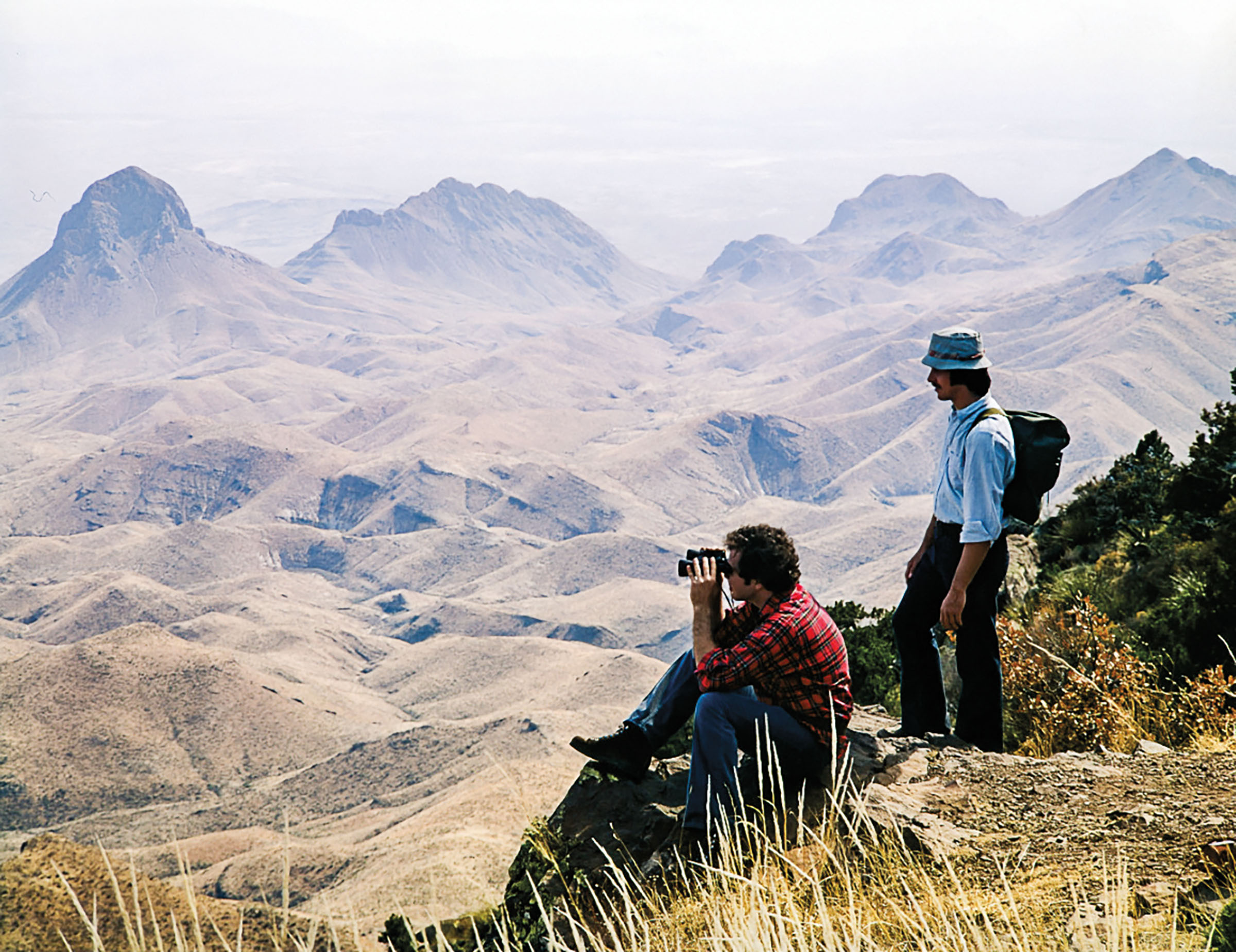 A pair of hikers stand on a bluff overlooking a valley and tall mountains