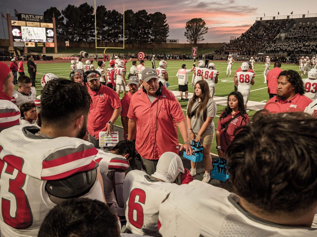 A man in a red jacket talks to a group of football players on a field at sunset