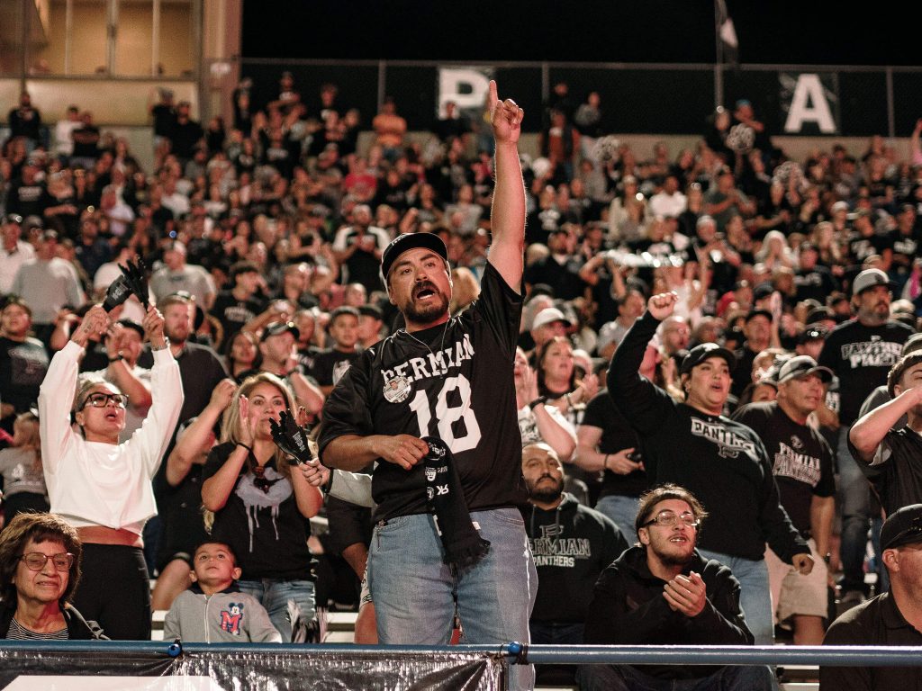 A man wearing a shirt reading "Permian / 18" stands cheering with his fist in the air in a crowd