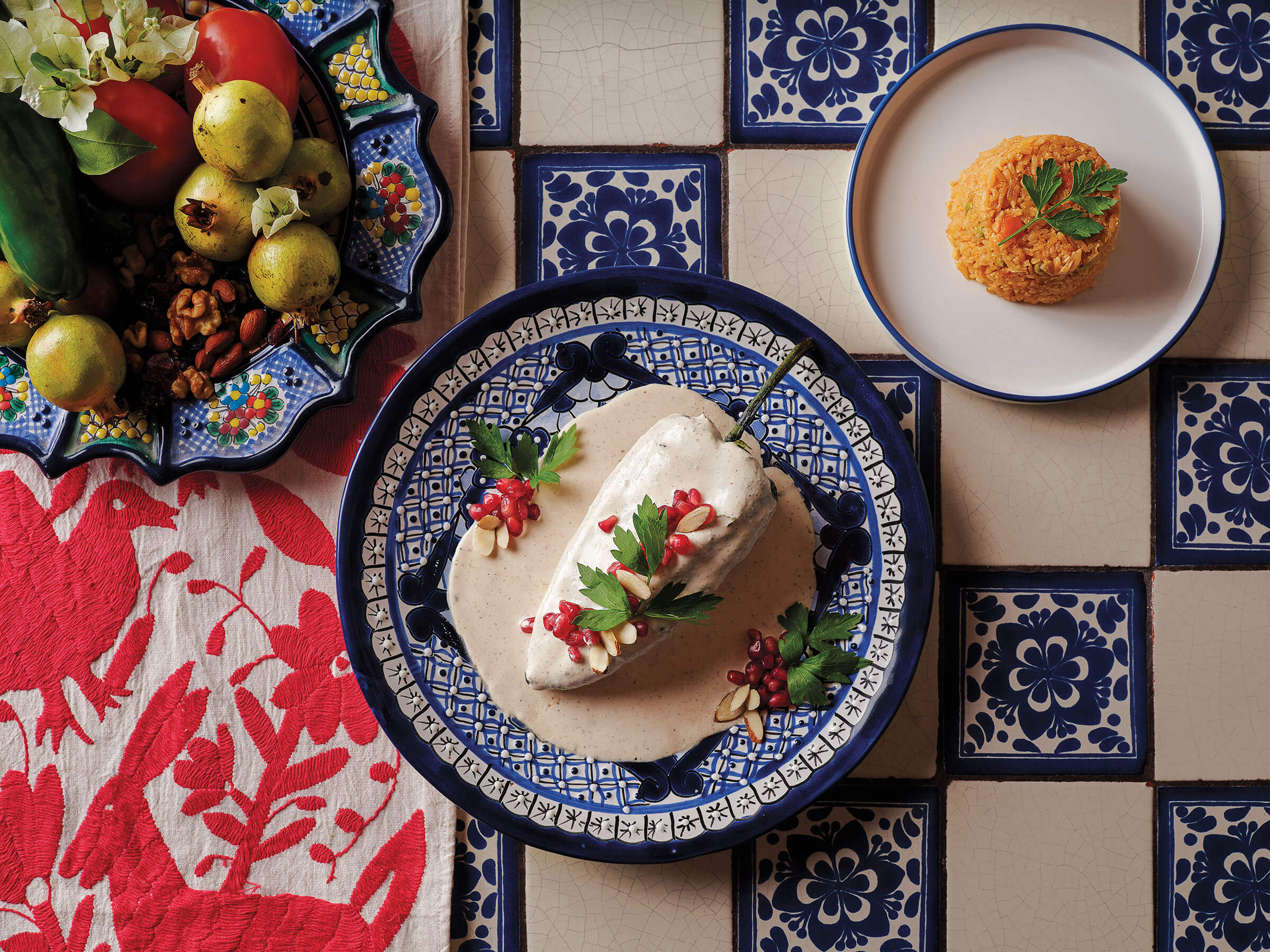 An overhead view of a chile in a white sauce with red pomegranate seeds. The food is plated on a blue plate on a blue and white checkered table