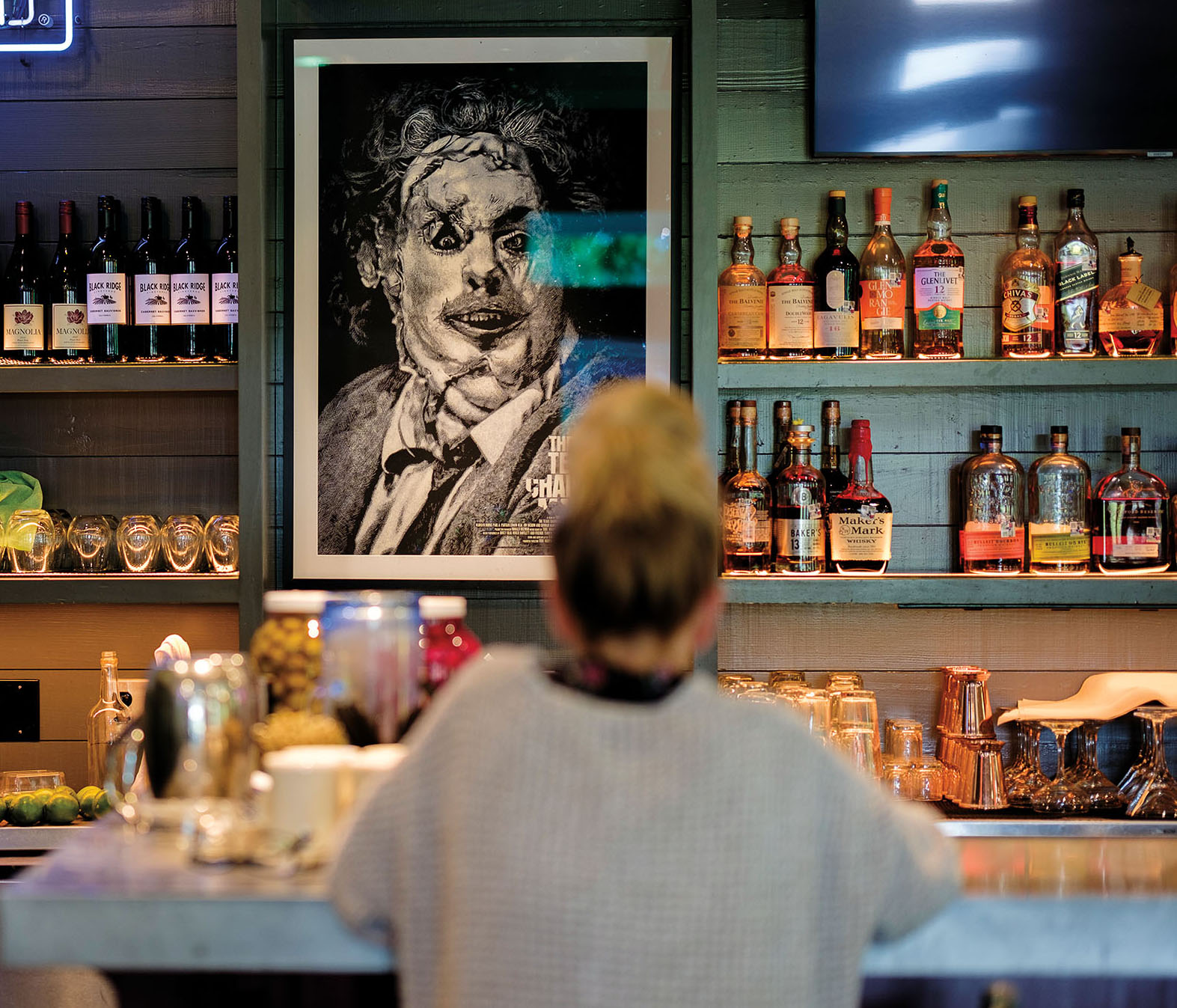 A woman sits at a bar in front of a black-and-white portrait of a scary face