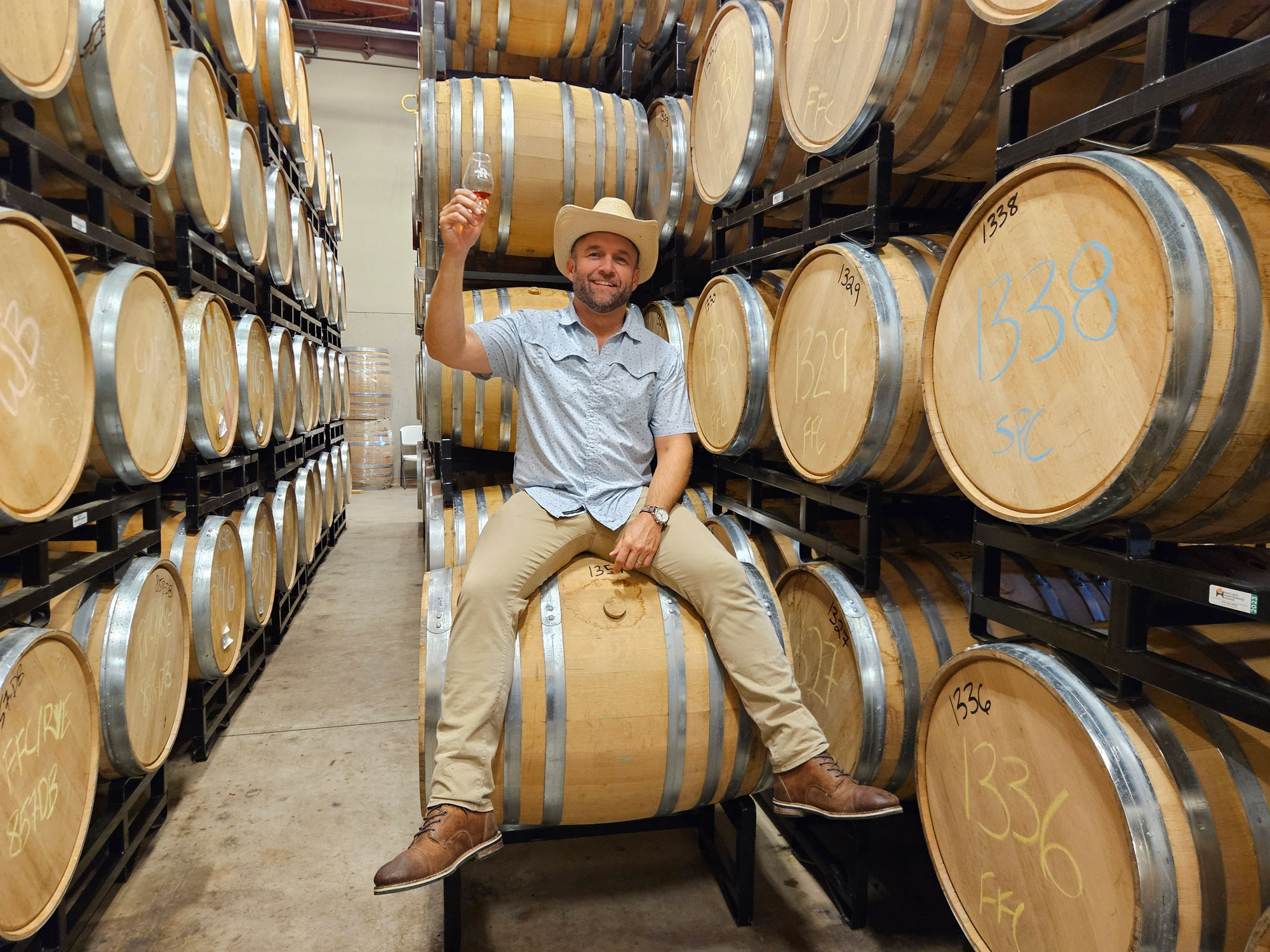 A man in a cowboy hat holding a glass of wine sits on a barrel in a distillery room