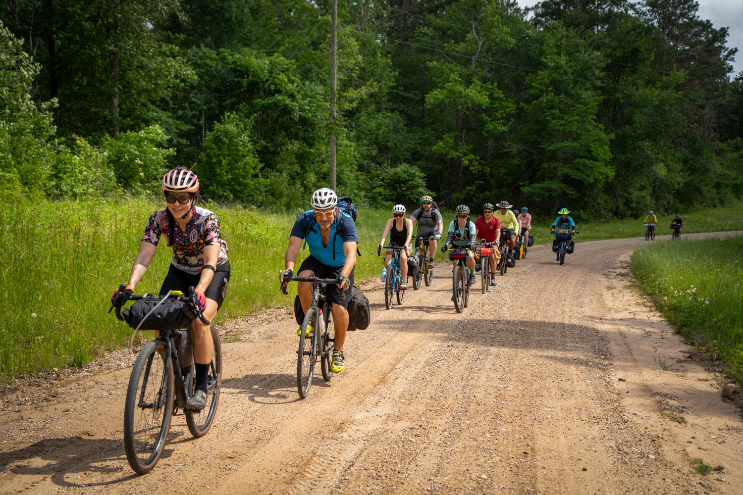 A group of several people on bikes wearing helmets and other bicycling gear ride down a gravel road surrounded by a green forest.