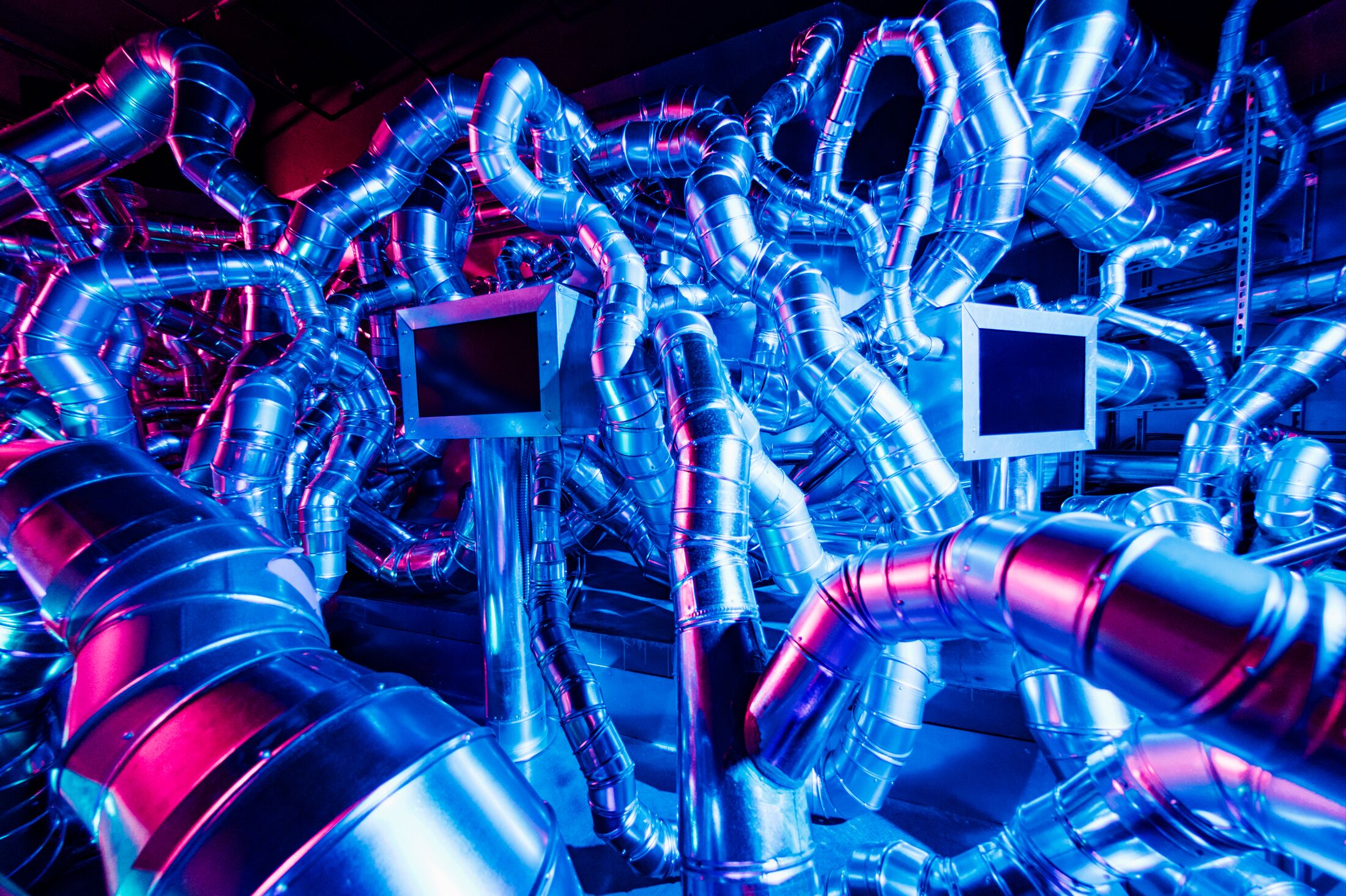 An image of silver pipes overlapping interspersed with black tv screens illuminated in a blue neon light