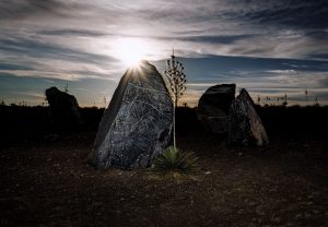 The sun shines behind two large stones in a desert setting