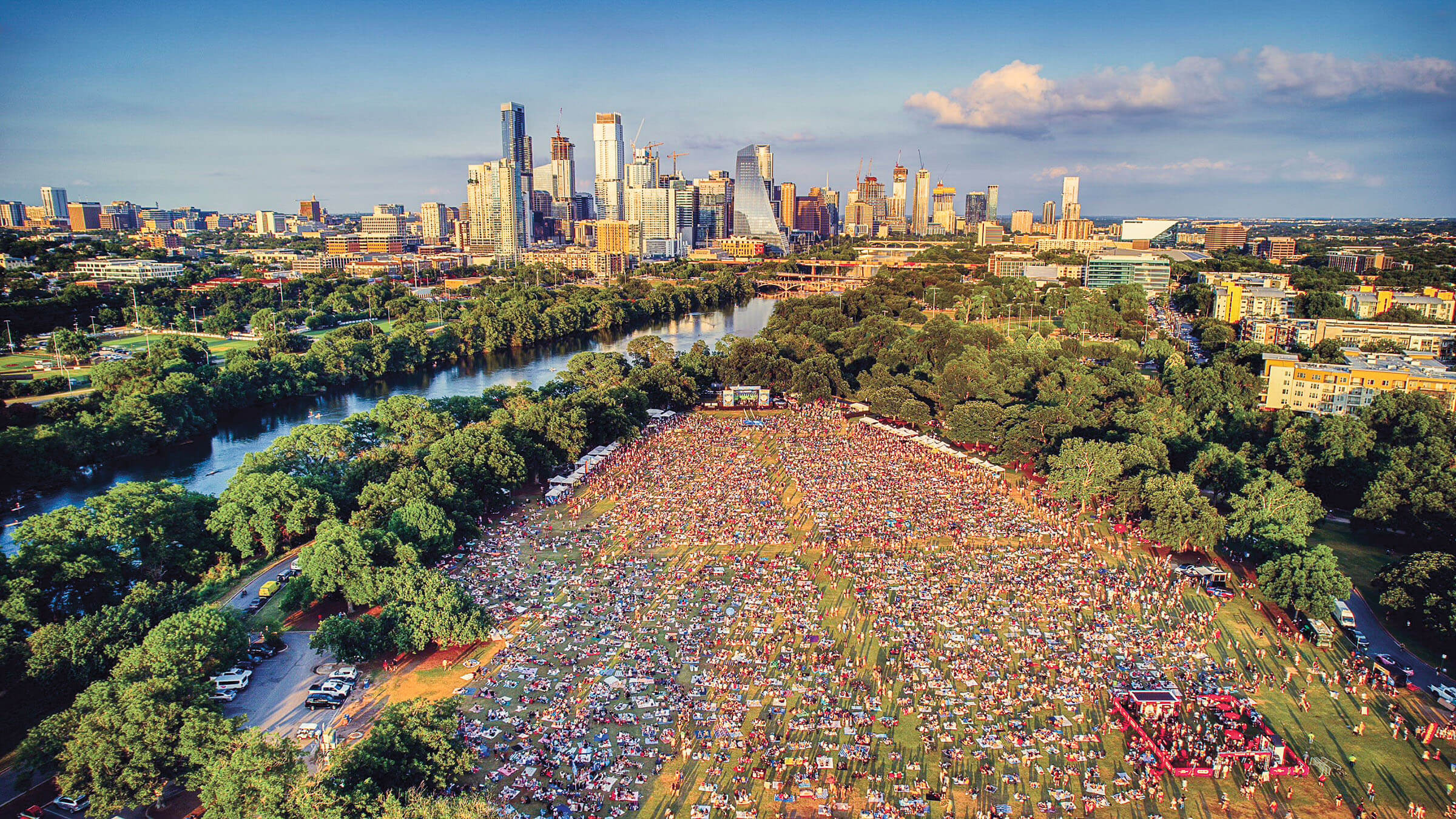 An overhead view of thousands of people on the grass in Zilker Park, with the Austin skyline and blue sky behind