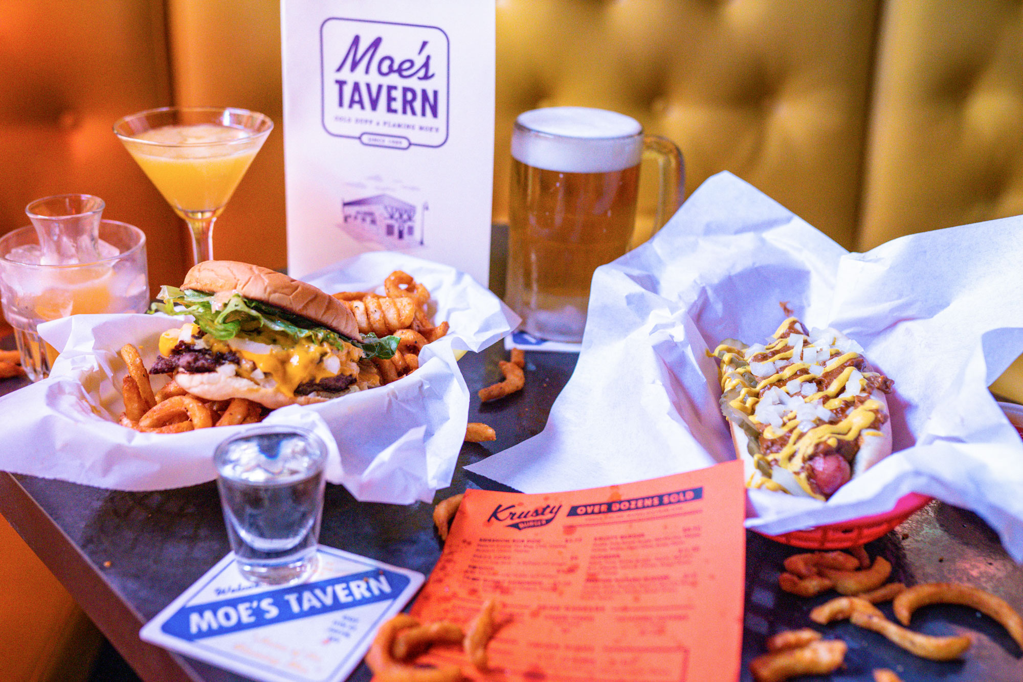 A basket with a burger and fries and another basket with a hot dog sit on a table surrounded by a cocktail and beer. Menus arranged around them read "Moe's Tavern."