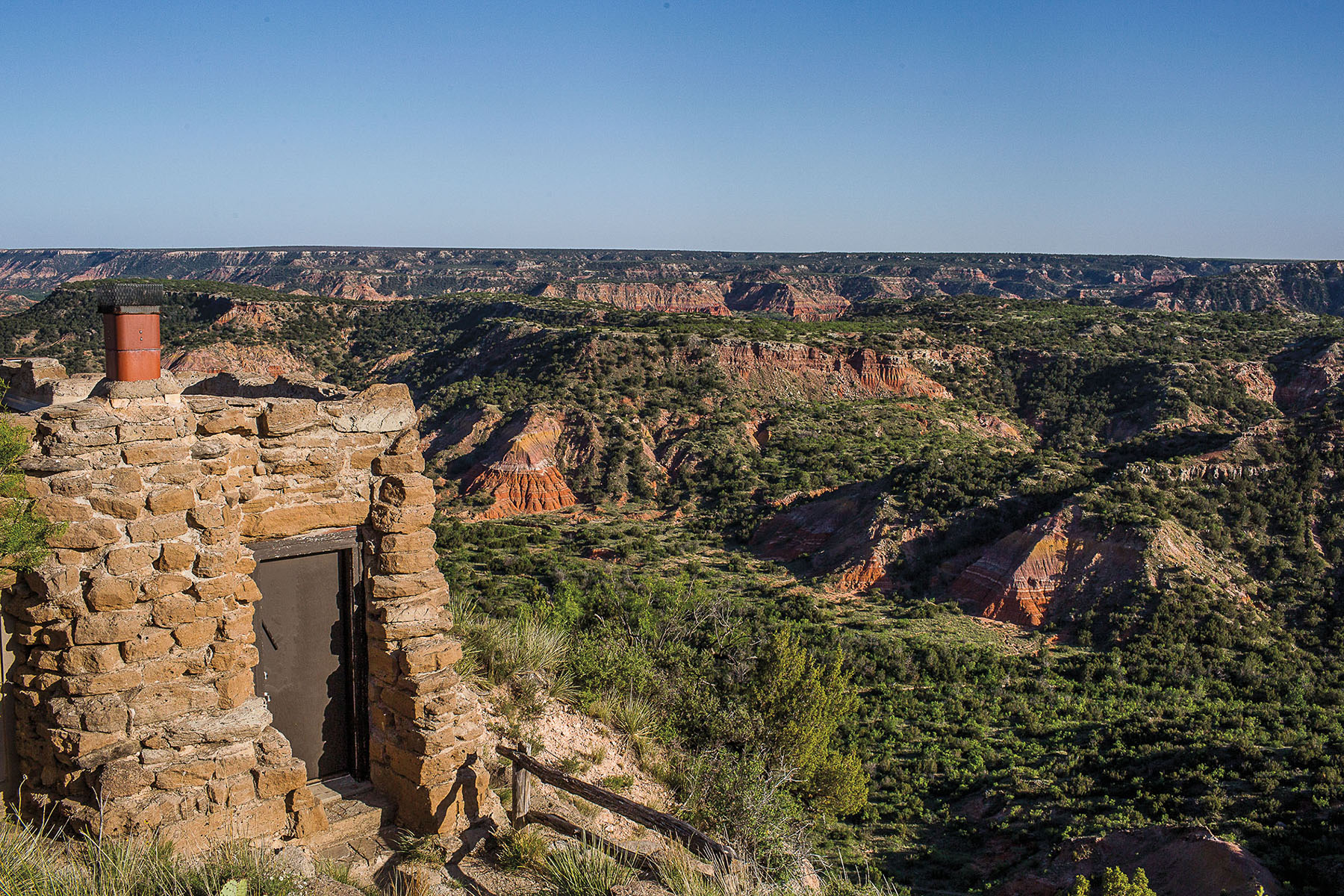 A stone cabin is built into the side of a red stone canyon. More of the canyon and trees are visible in the distance.