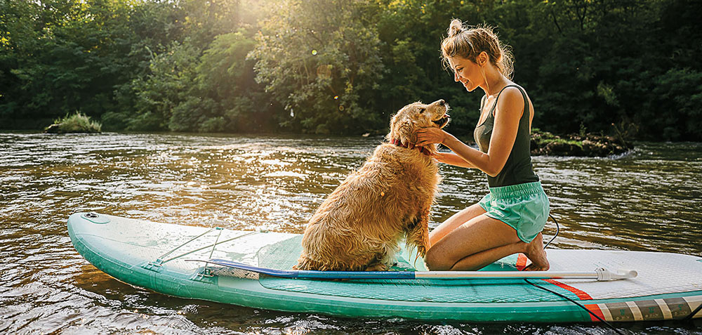 A woman kneels on a turquoise paddle board, petting a dog while floating on a body of water 