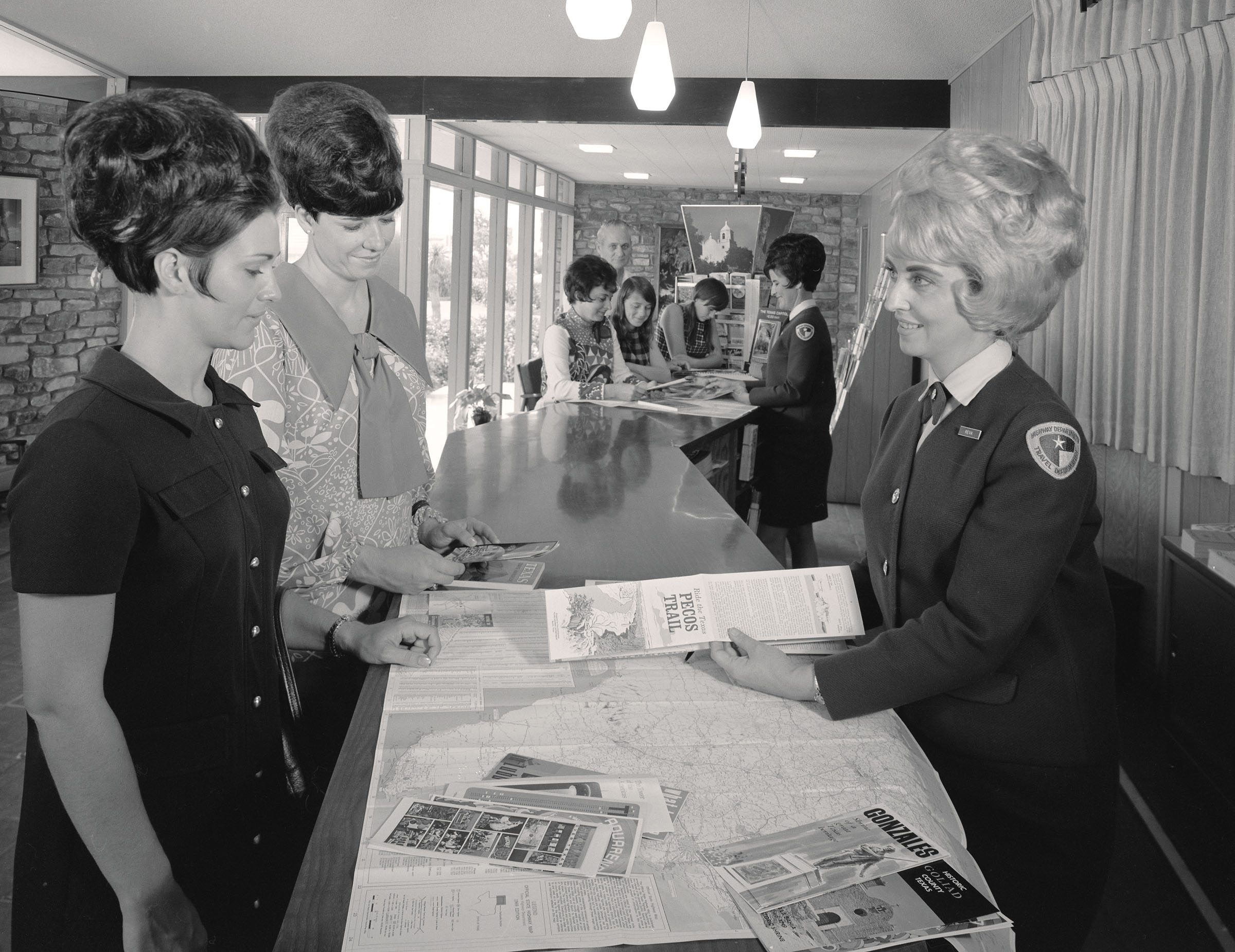A woman wearing an official-looking uniform holds a large brochure reading 'Pecos Trail' over a large map of Texas. Two other women, on the other side of the counter, smile as they look at the information