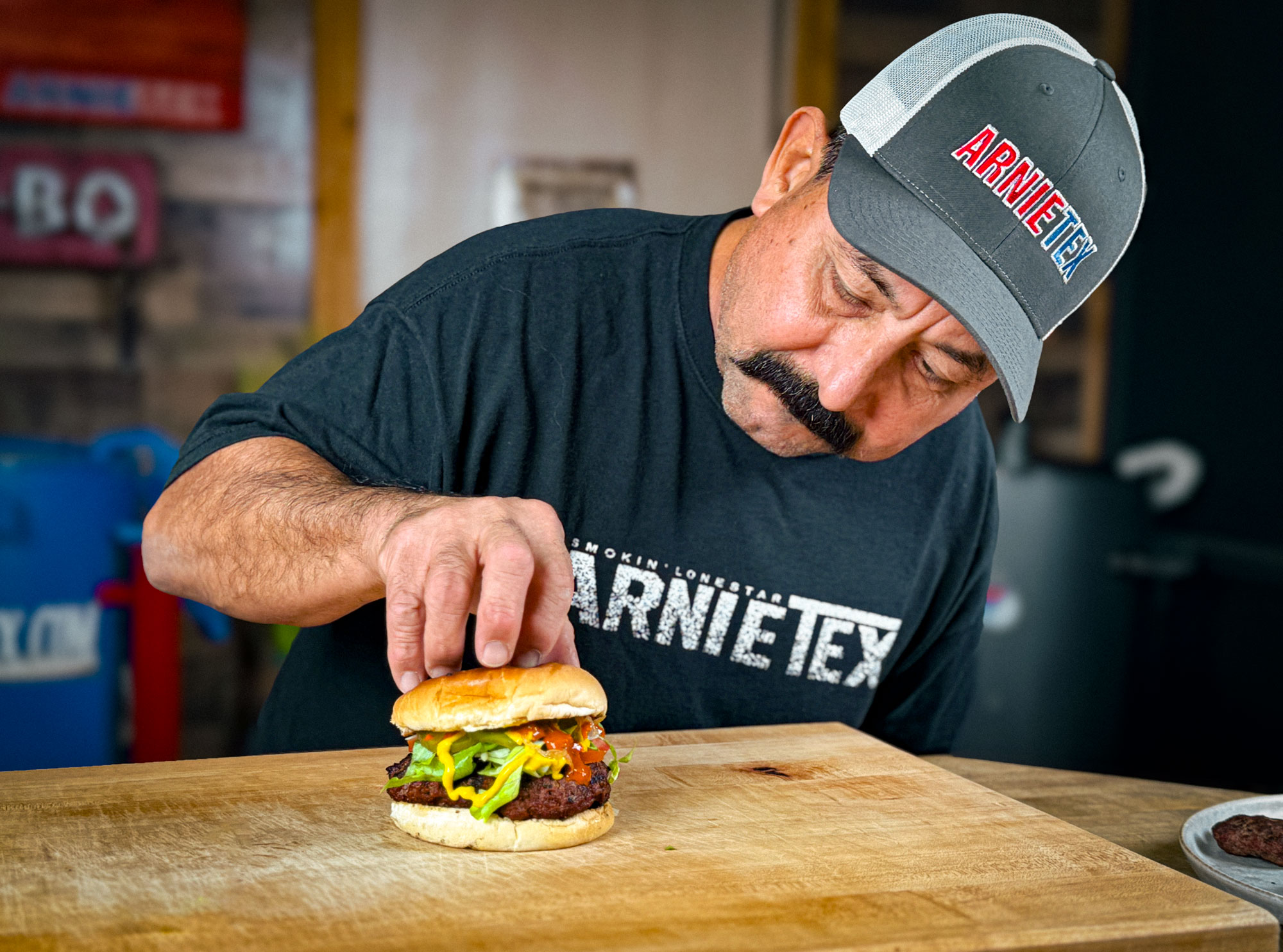 A man in a cap and tshirt bends over a table with a cutting board while touching a burger with meat, lettuce, and tomato.
