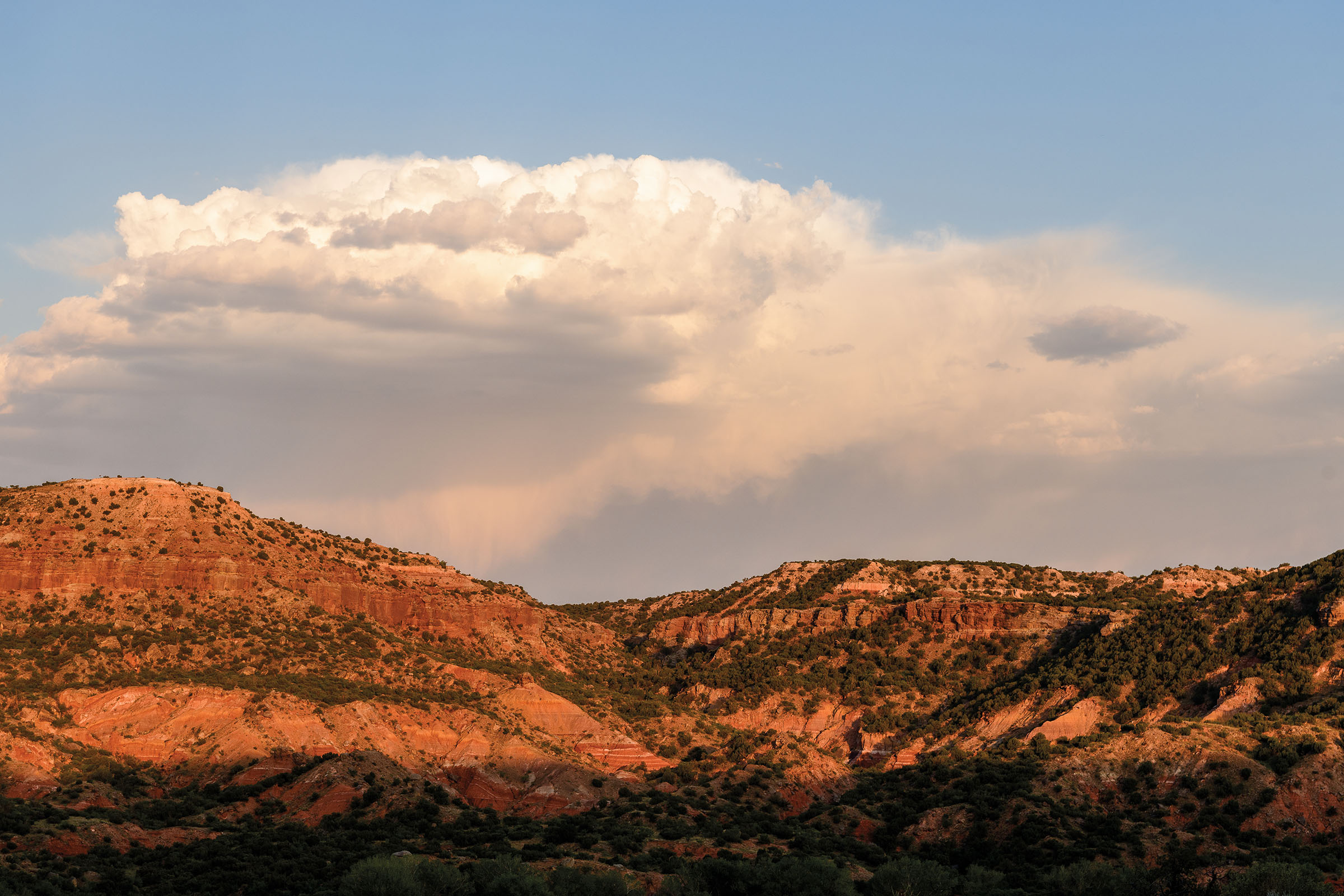 Large clouds build over a wide rocky landscape dotted with green scrub
