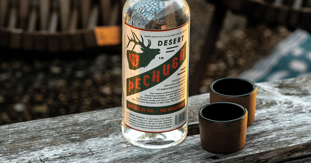 A bottle of clear liquor and two cups on a wooden bench