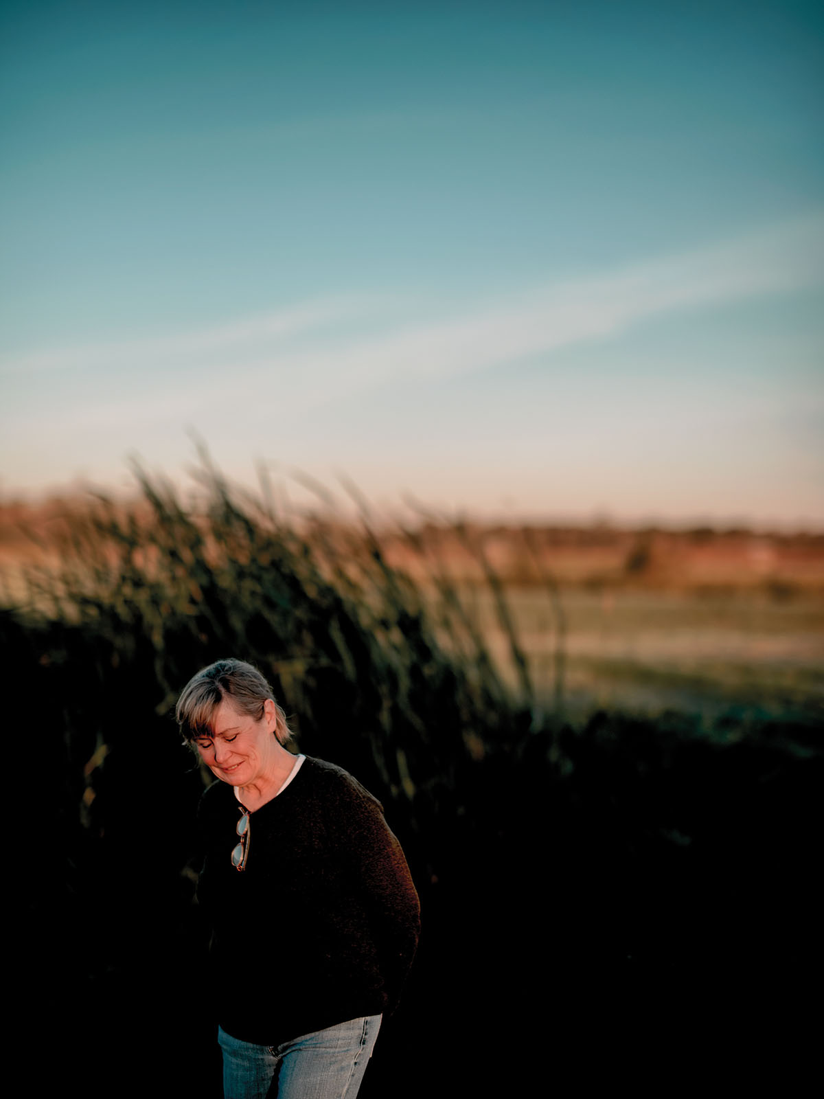 A person walks with their head bowed in front of tall grass and a sunset