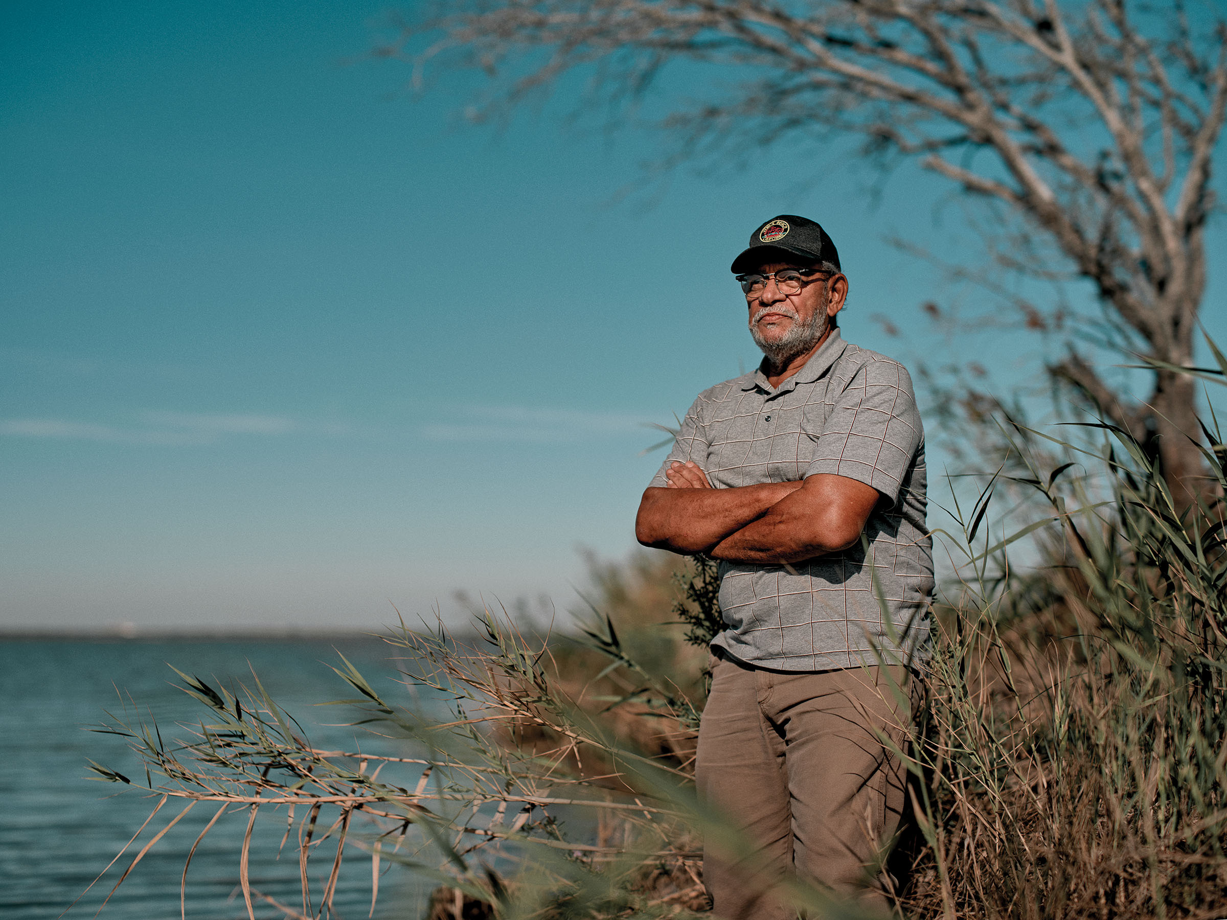 A man in a baseball cap stands with his arms crossed in front of a marshy area