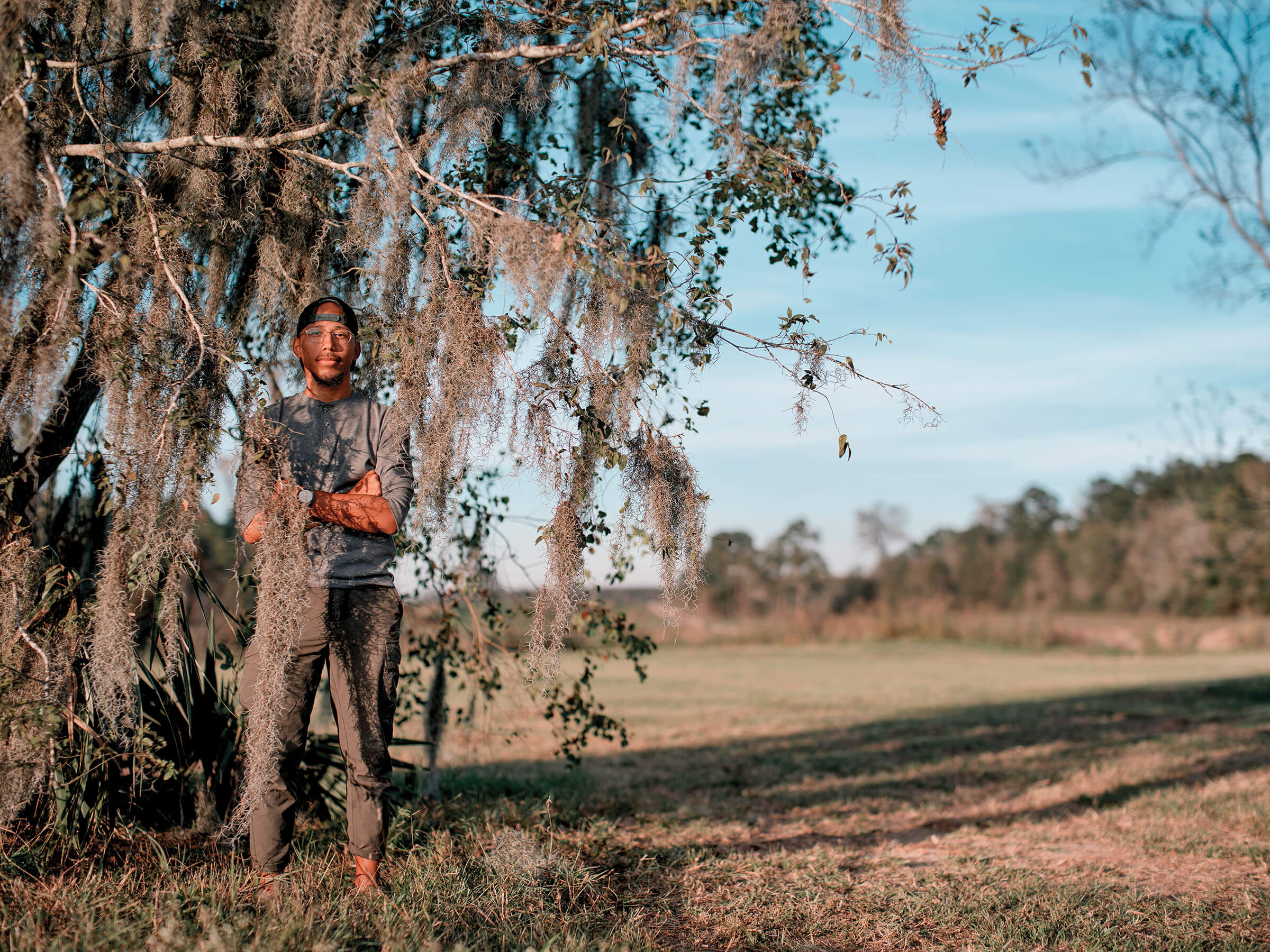 A man in a gray shirt stands with his arms crossed next to a tree covered in moss