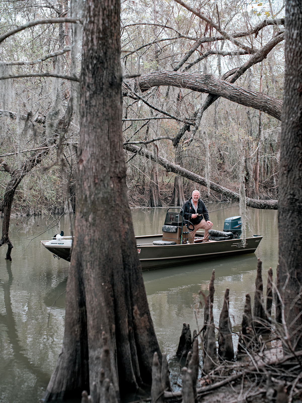 A man poses from a gray metal boat in murky water beneath tall trees
