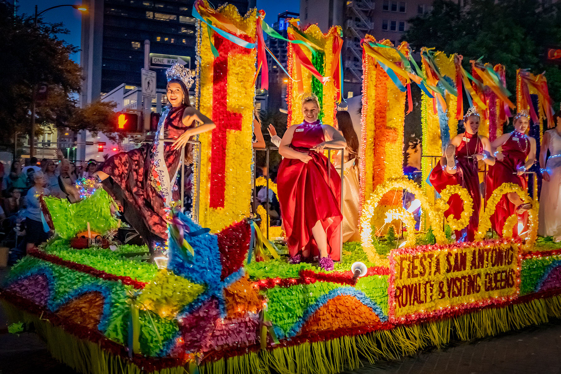 Women in colorful dresses stand on a vibrant, multicolored float that says FIESTA.