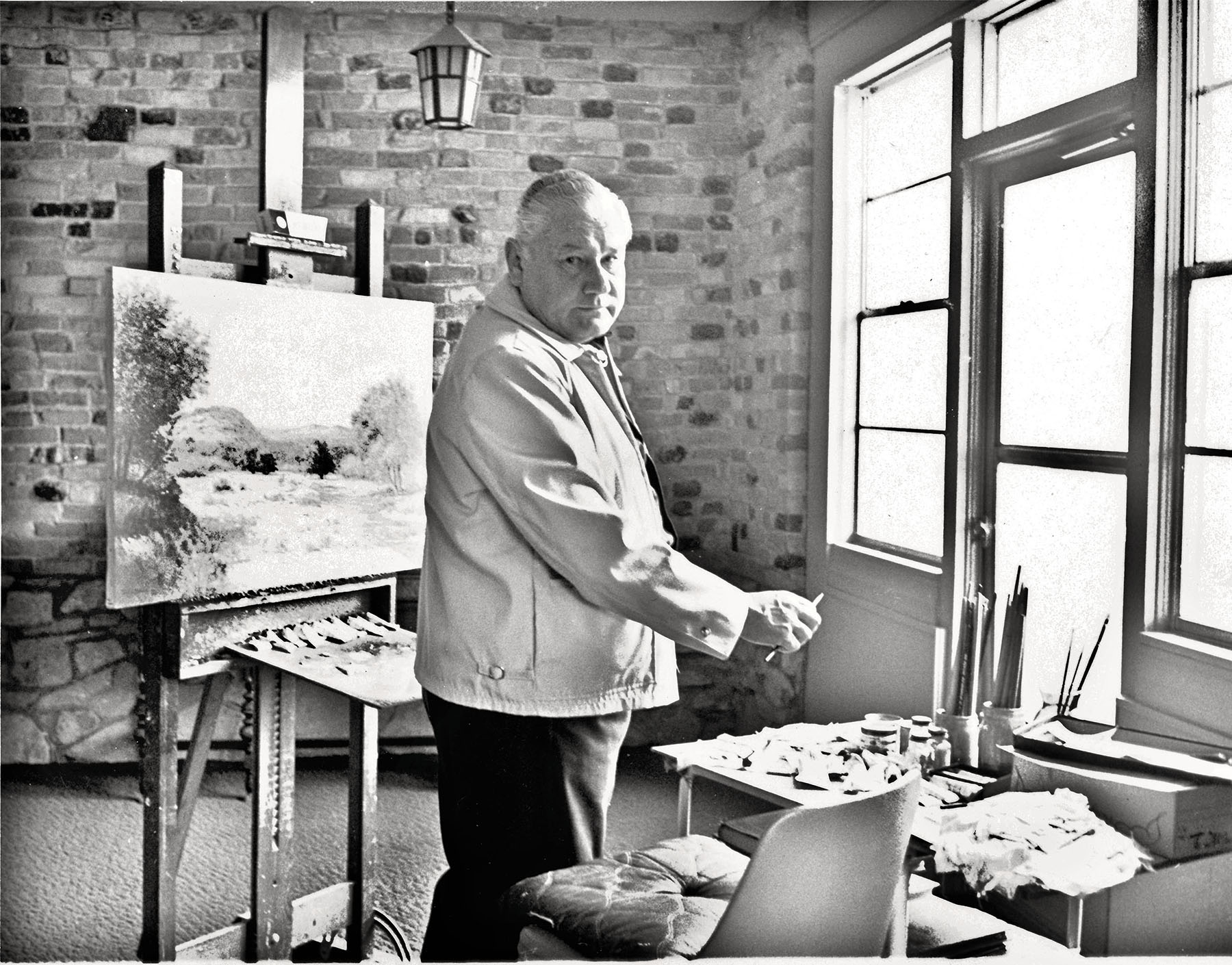 A black and white photograph of a man painting in a studio with large windows and a canvas on display
