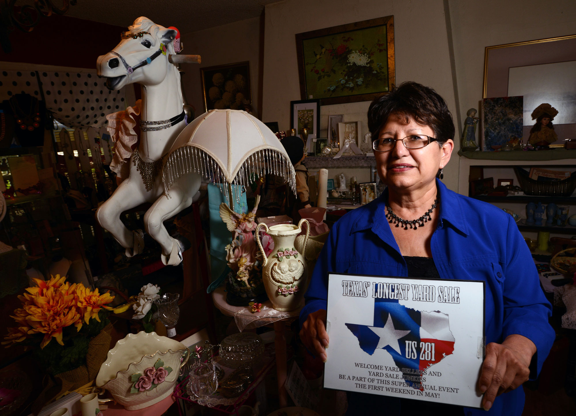 A woman holds a sign promoting Texas' Longest Yard Sale sits in a room full of antiques.