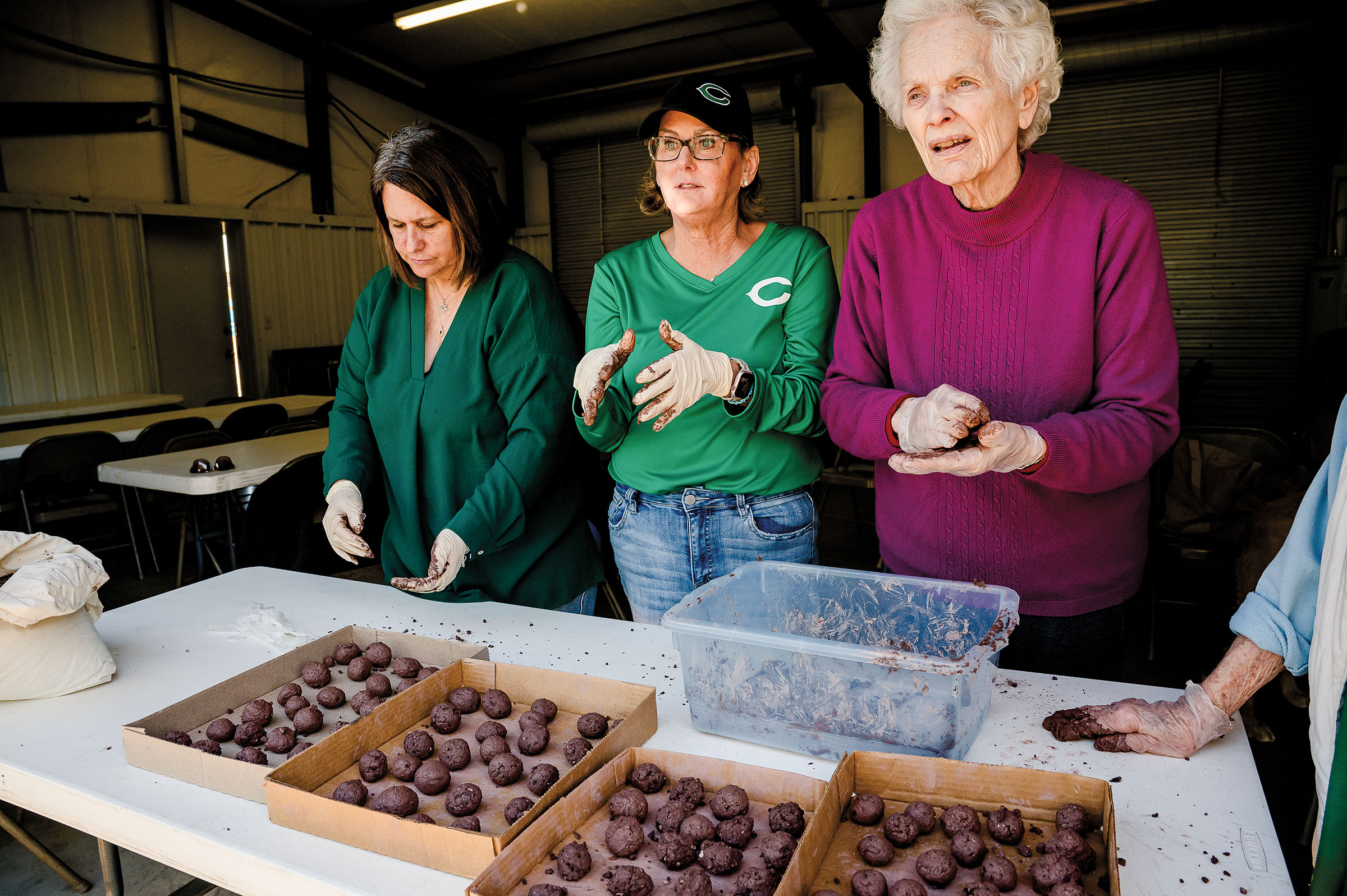 Three people stand at a plastic table creating brown balls of dirt and seeds