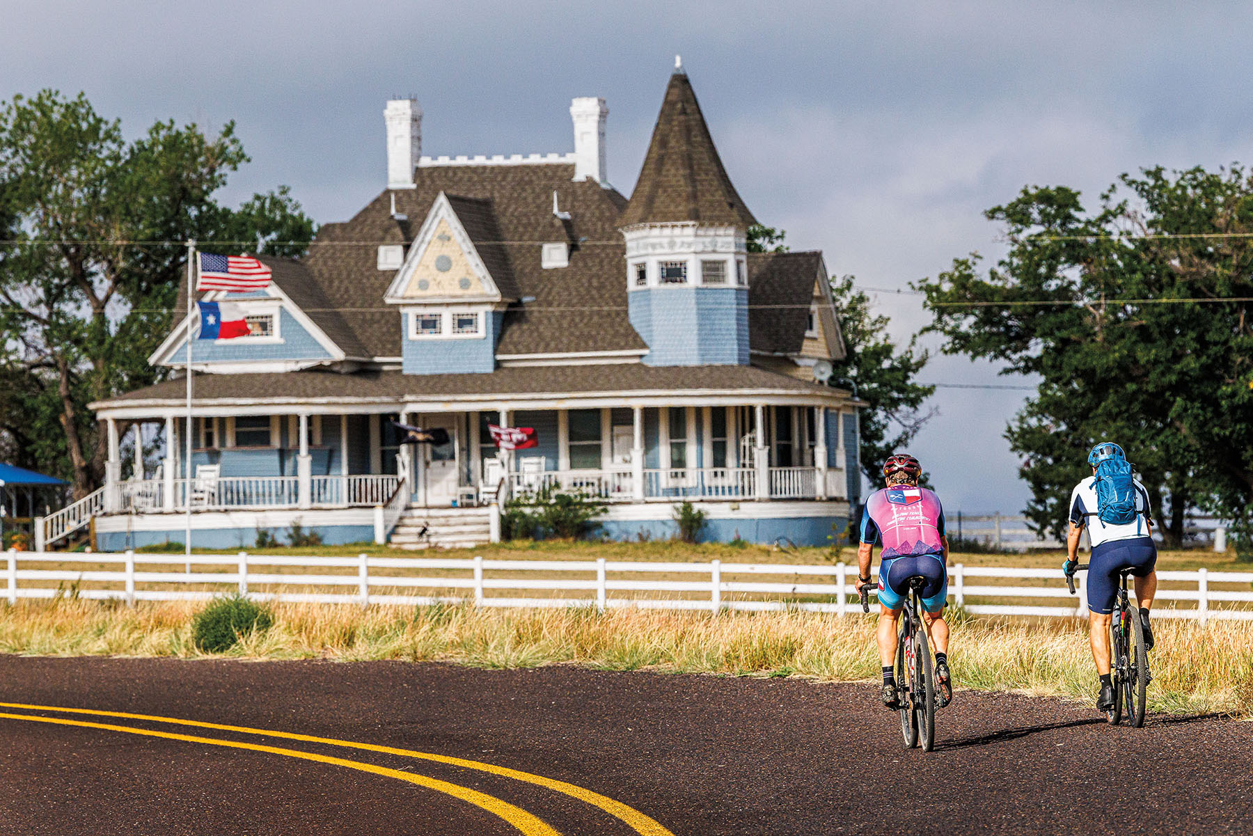 Two people bike next to a large blue Victorian home in a grassy field