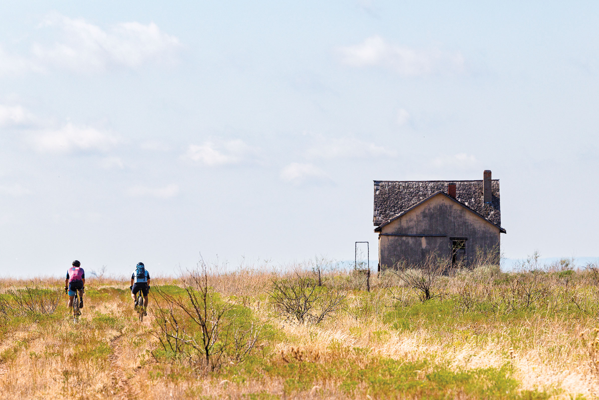 Two people ride bicycles next to an old wooden building in a field