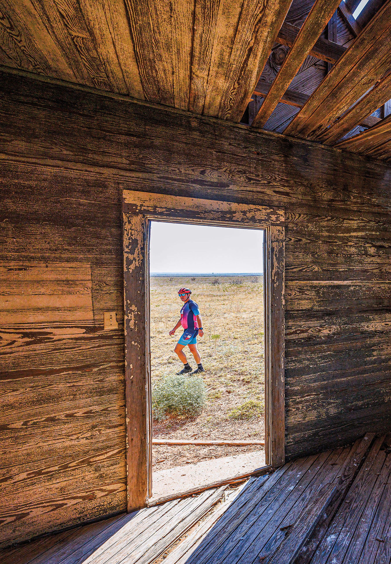 A person wearing brightly-colored bicycle outfit and helmet stands in the doorway of a wooden-frame building