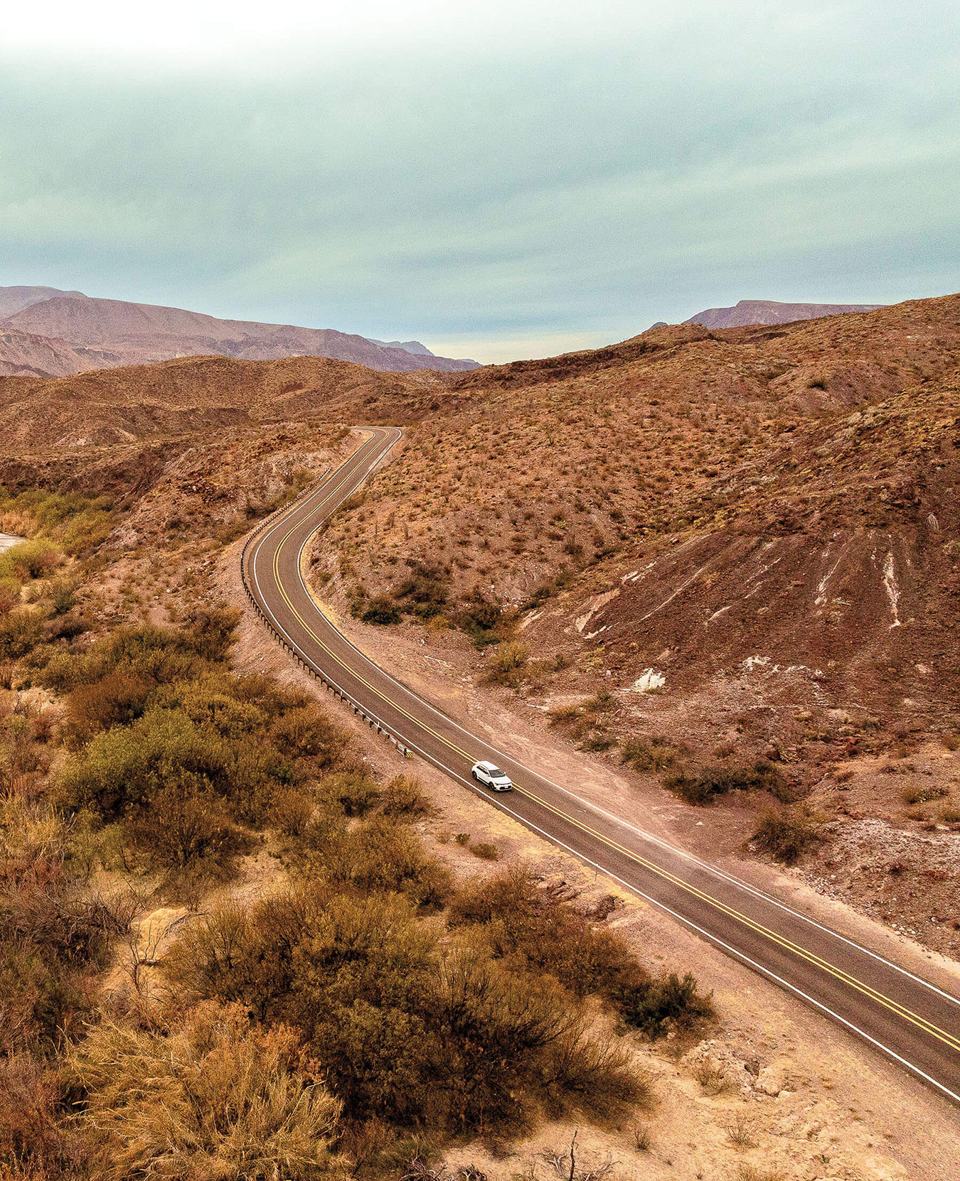 A white electric vehicle drives along a dark asphalt road in a desert landscape next to the Rio Grande