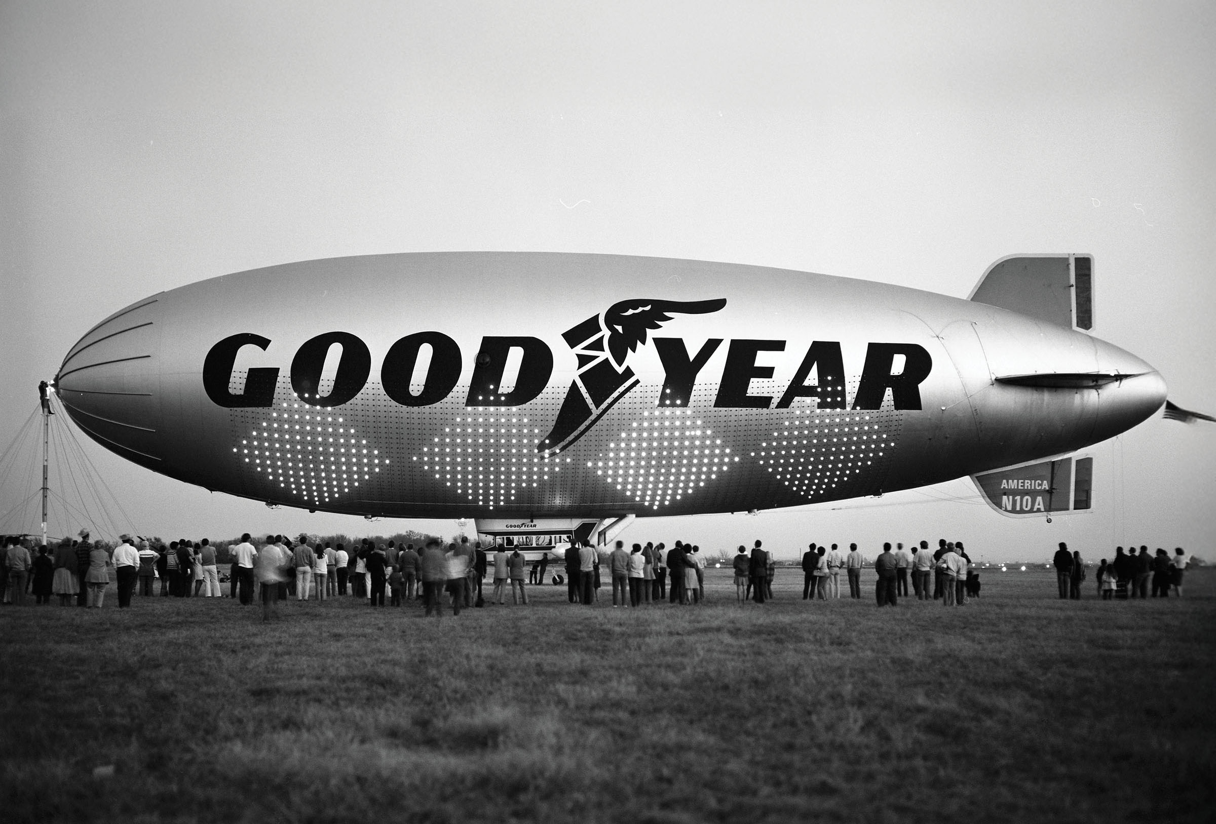 A group of people gather underneath a large silver blimp reading 'Goodyear'