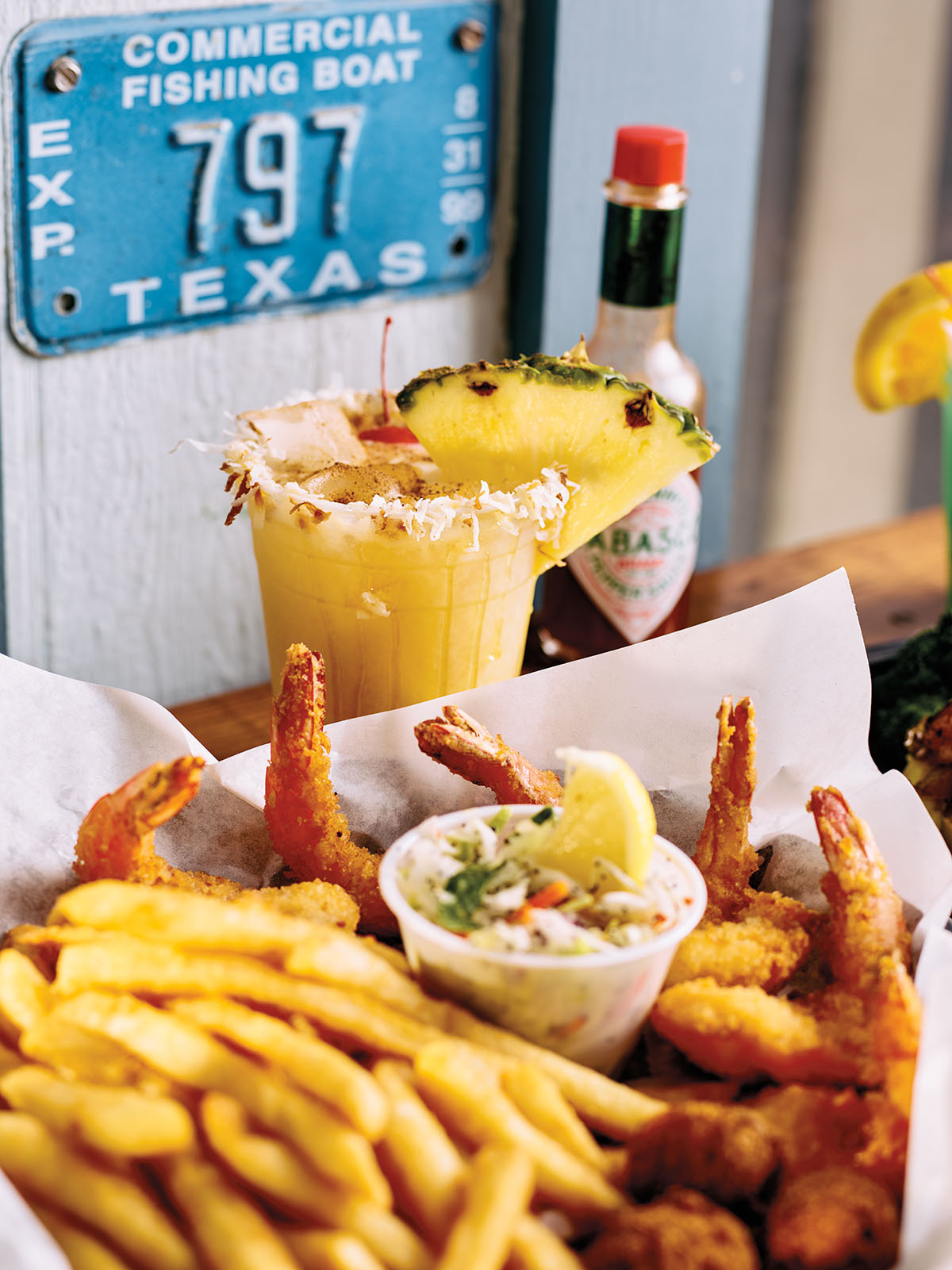 A golden cocktail with a large pineapple garnish next to a basket lined with paper filled with fries, shrimp, and coleslaw