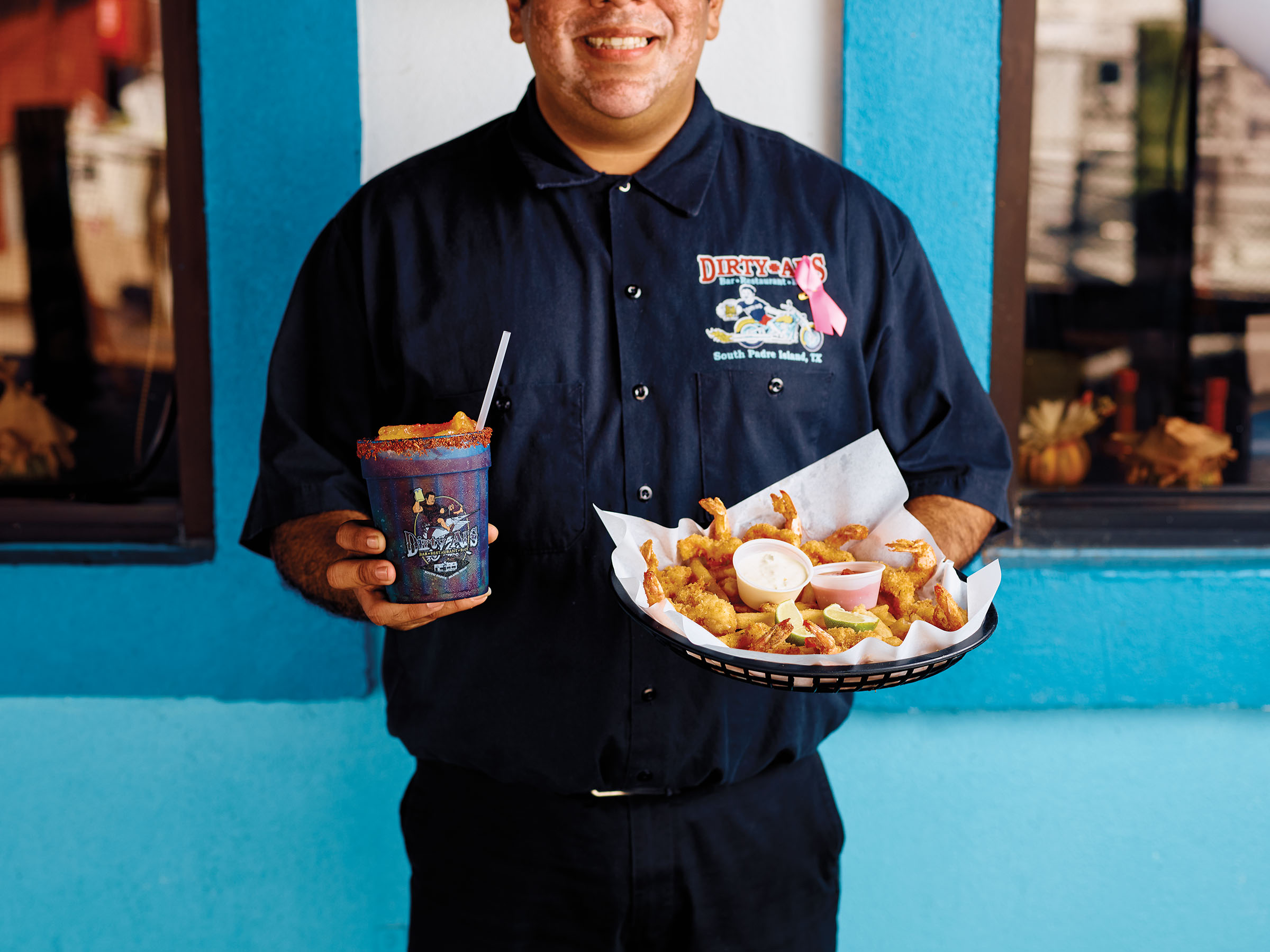 A person wearing a work uniform reading "Dirty Al's" holds a plastic cup full of margarita and a large platter of golden fish