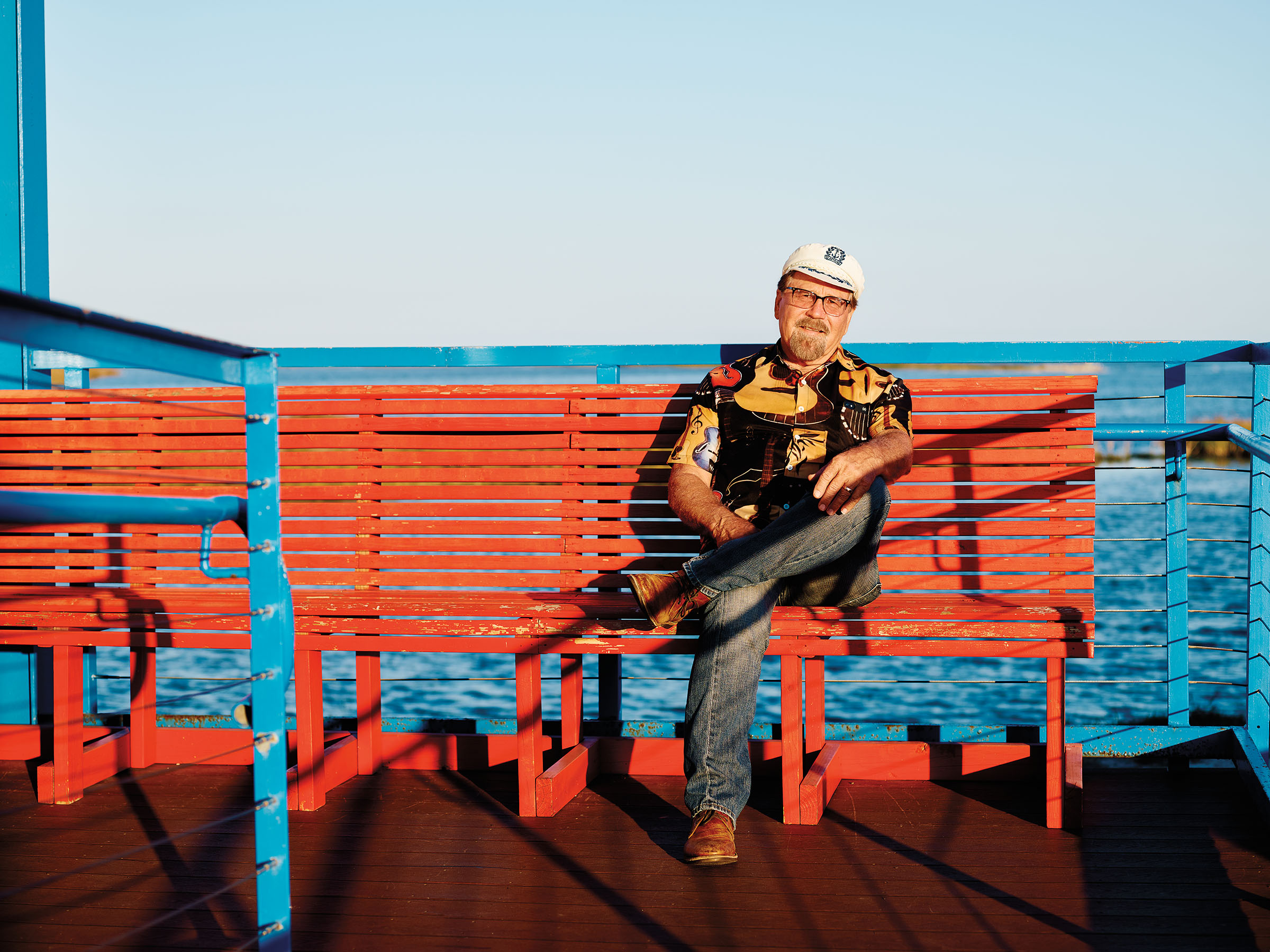 A man in a sailor's cap sits on a bright red bench in front of blue water