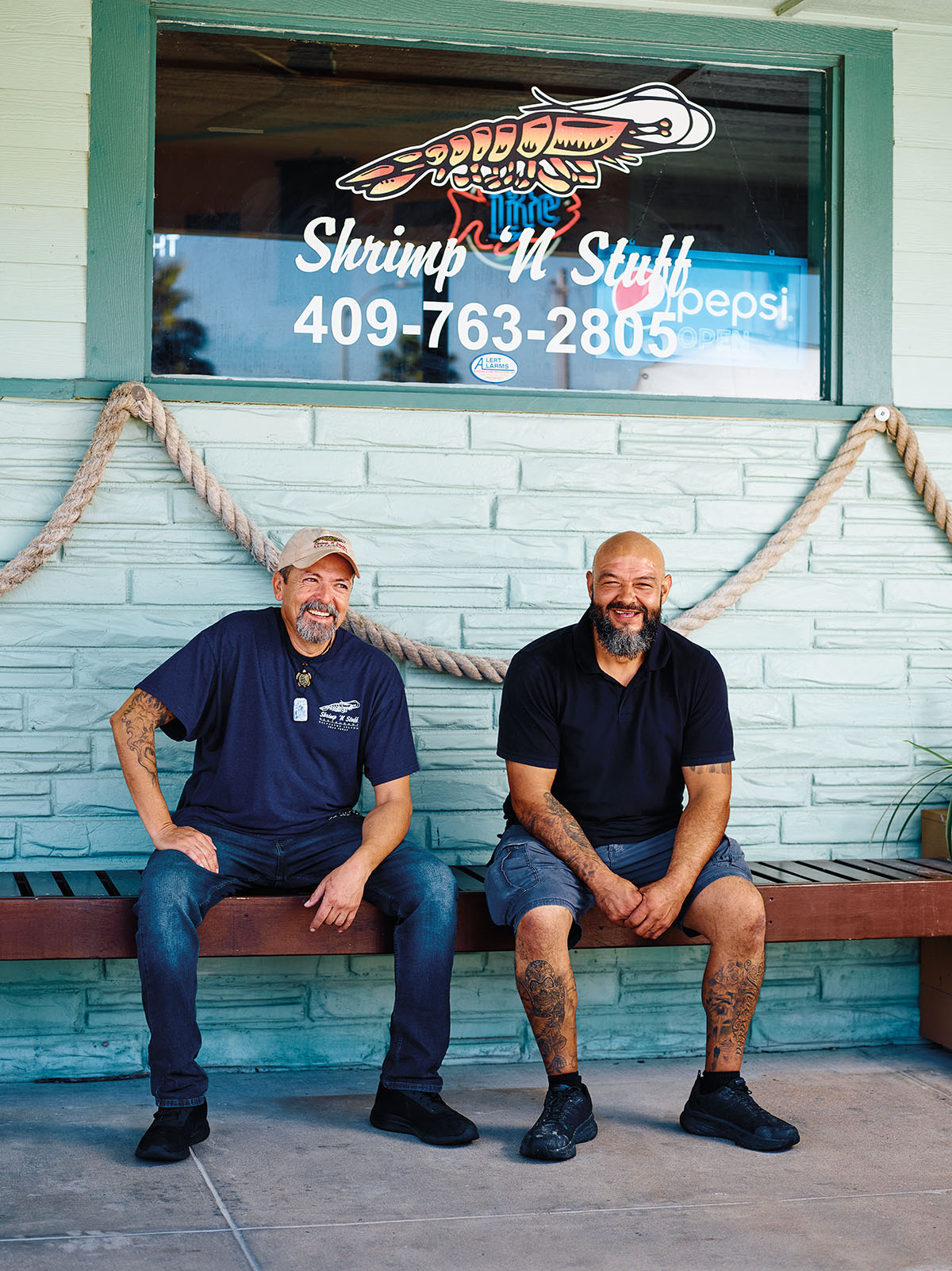 Two men sit on a wooden bench in front of a blue-painted brick wall. A window above them reads 'Shrimp n Stuff'