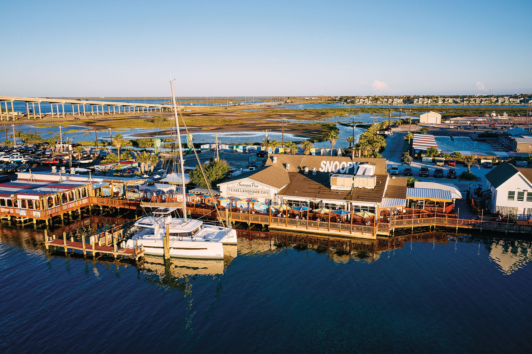 An overhead drone photograph of a long pier with a white building and tall white sailboat parked in front. Numerous tables with umbrellas line the pier