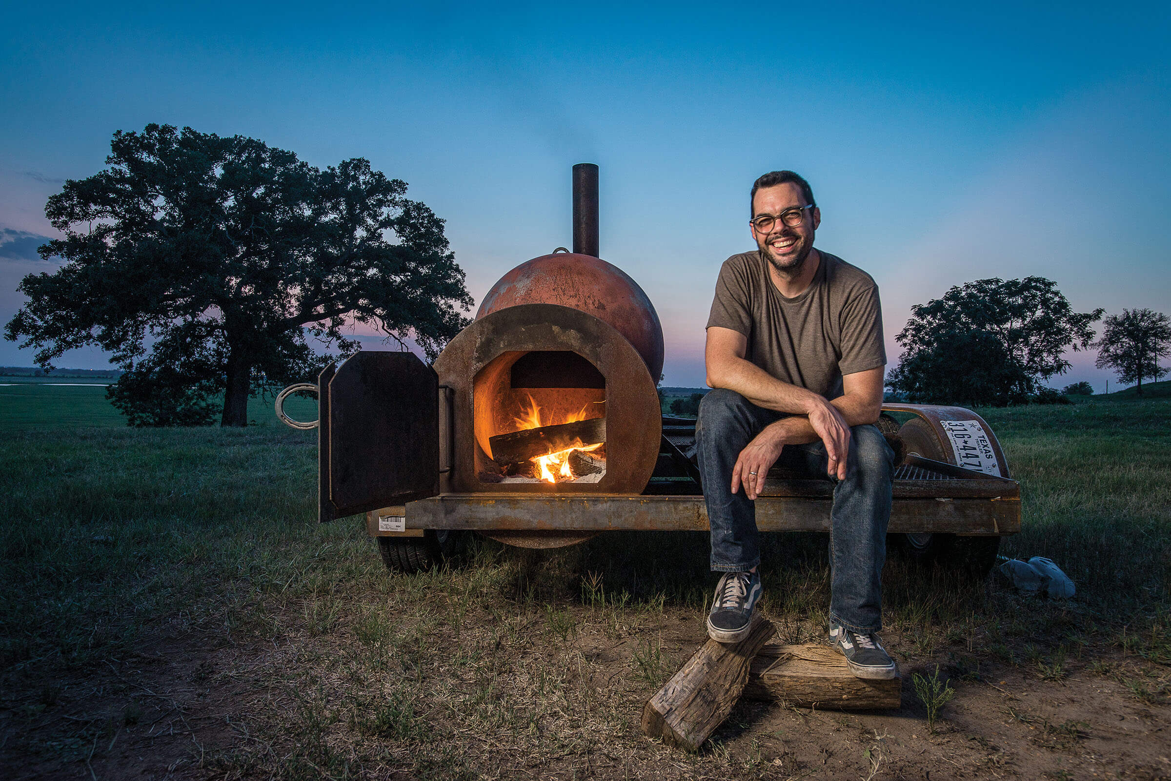 A man in a t-shirt sits next to a pit smoker where fire is visible. The scene is an outdoor setting at dusk.