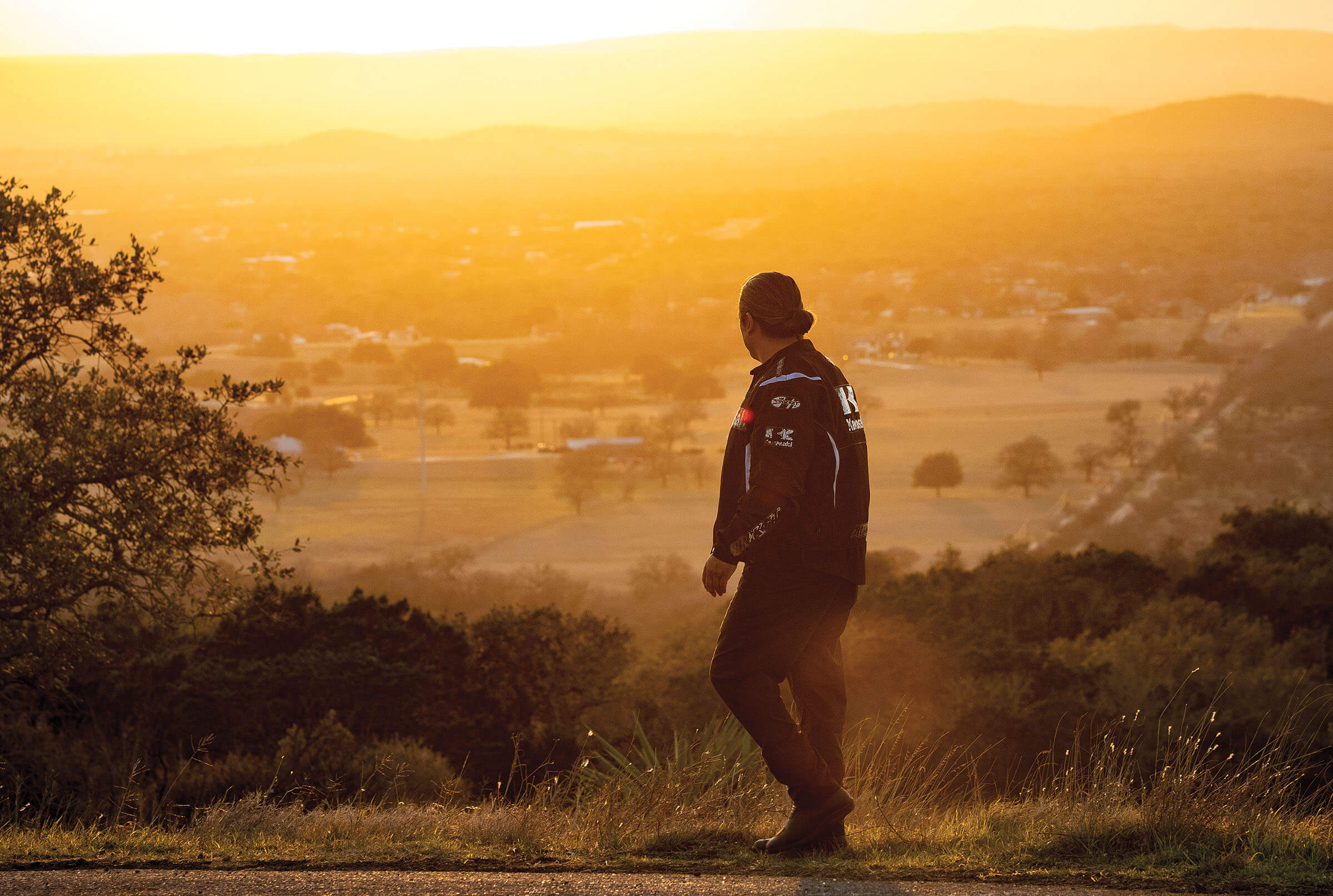 A man in a motorcycle jacket looks over a grassy overlook to the valley below in golden sunset light