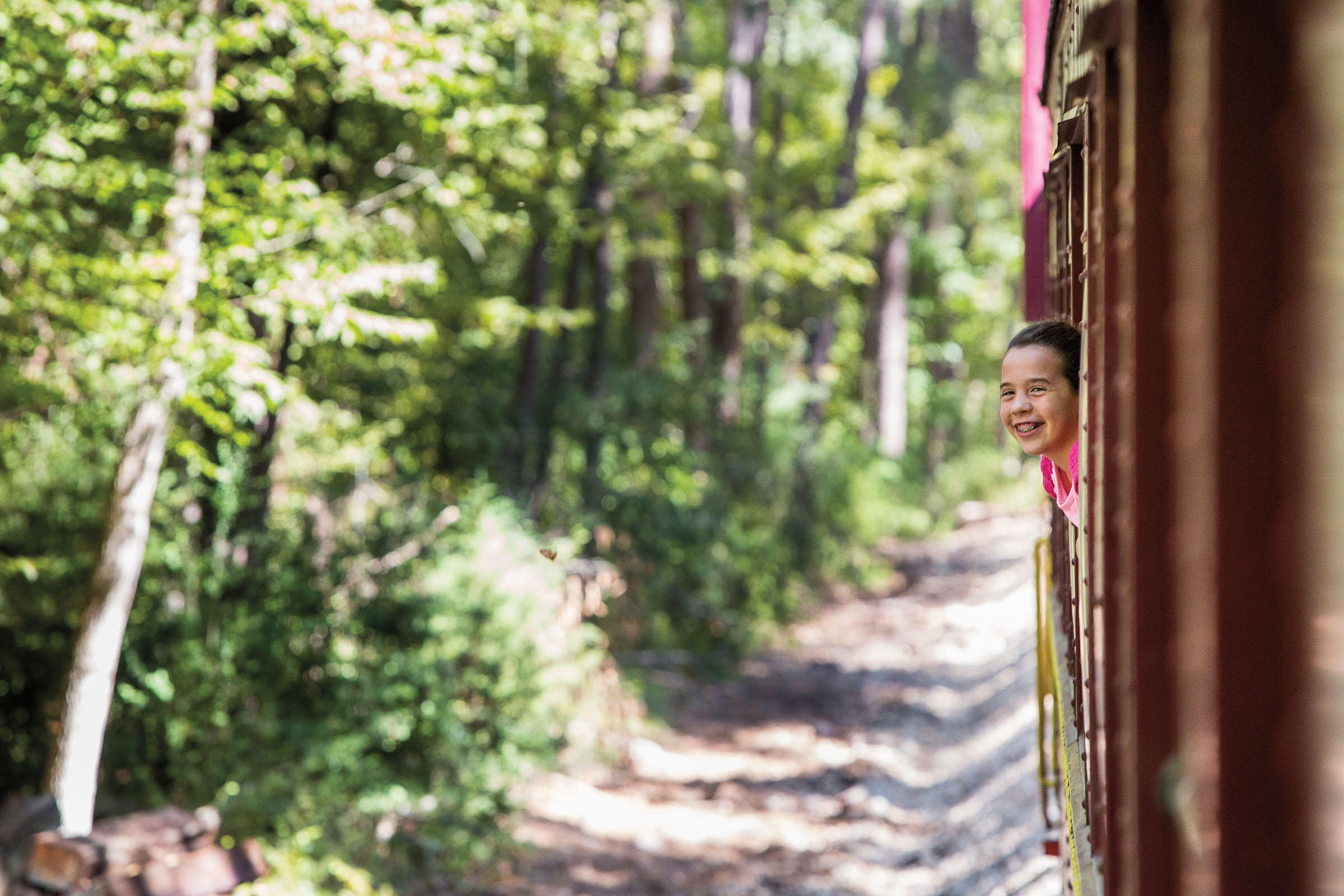 A young person smiles while leaning her head out of the window of a red train car in a natural setting