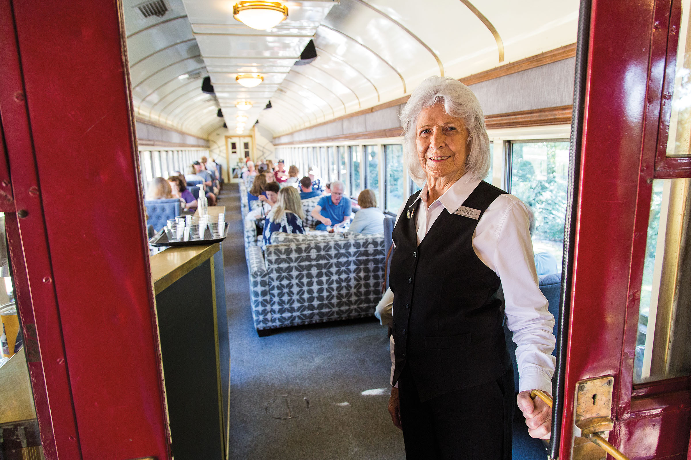 A woman wearing a vest uniform holds open the door to an ornate train car