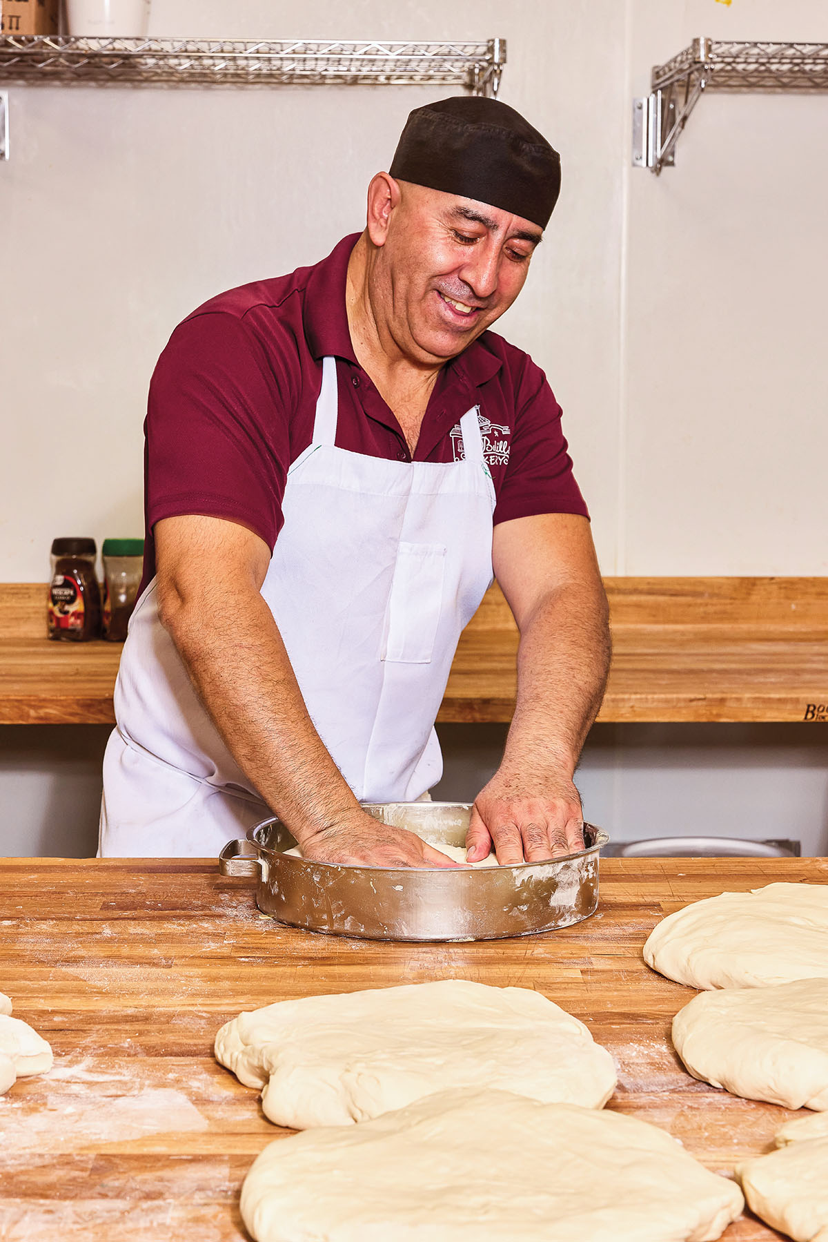 A man wearing a white apron and hat kneads dough on a wooden surface