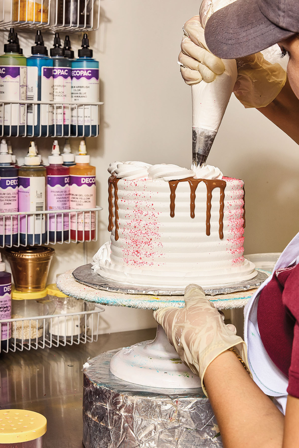 A person pipes frosting on a tall cake with a chocolate drip down the side