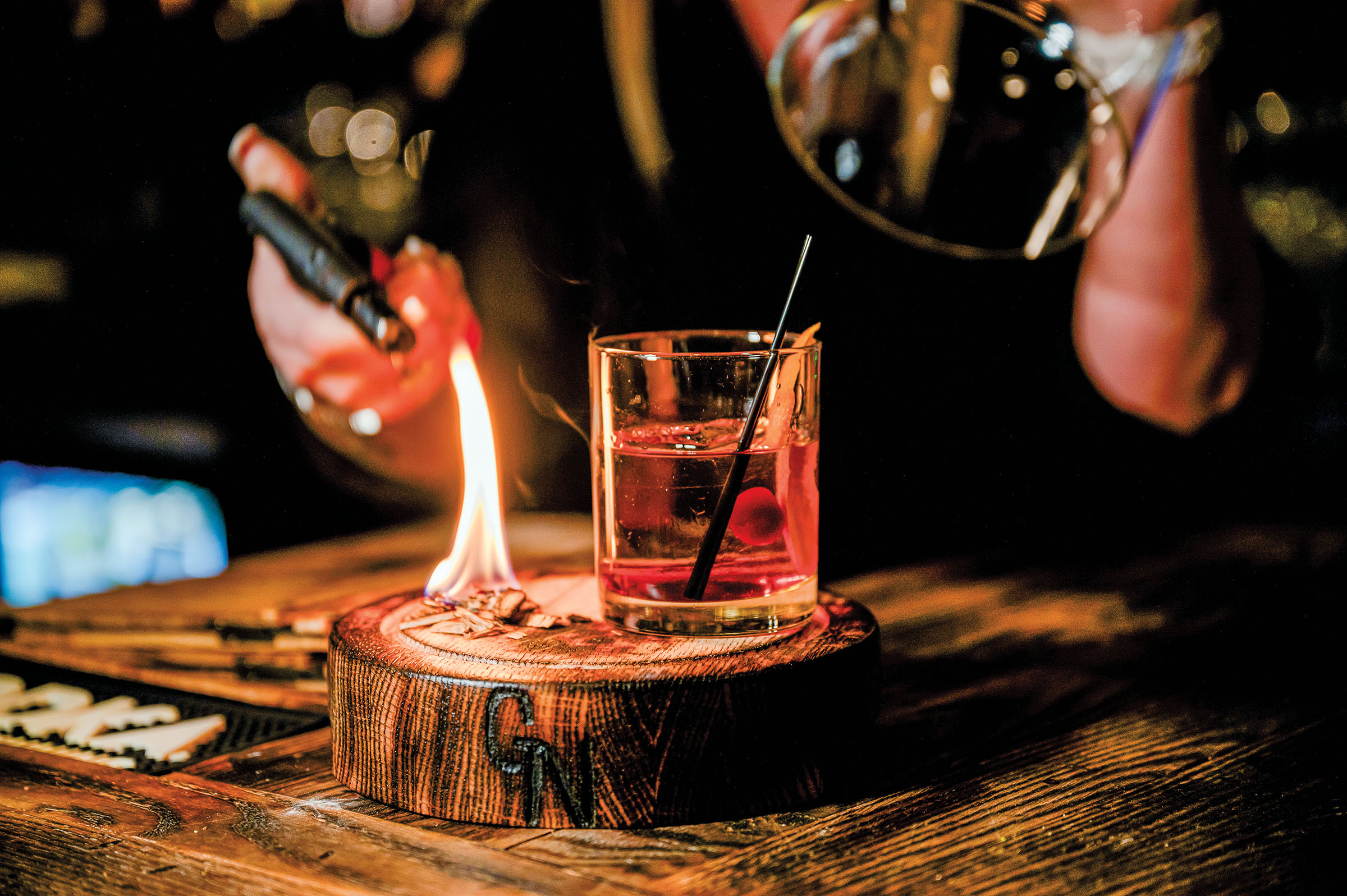 A person uses a small torch to light a flame next to a red cocktail on a wooden board