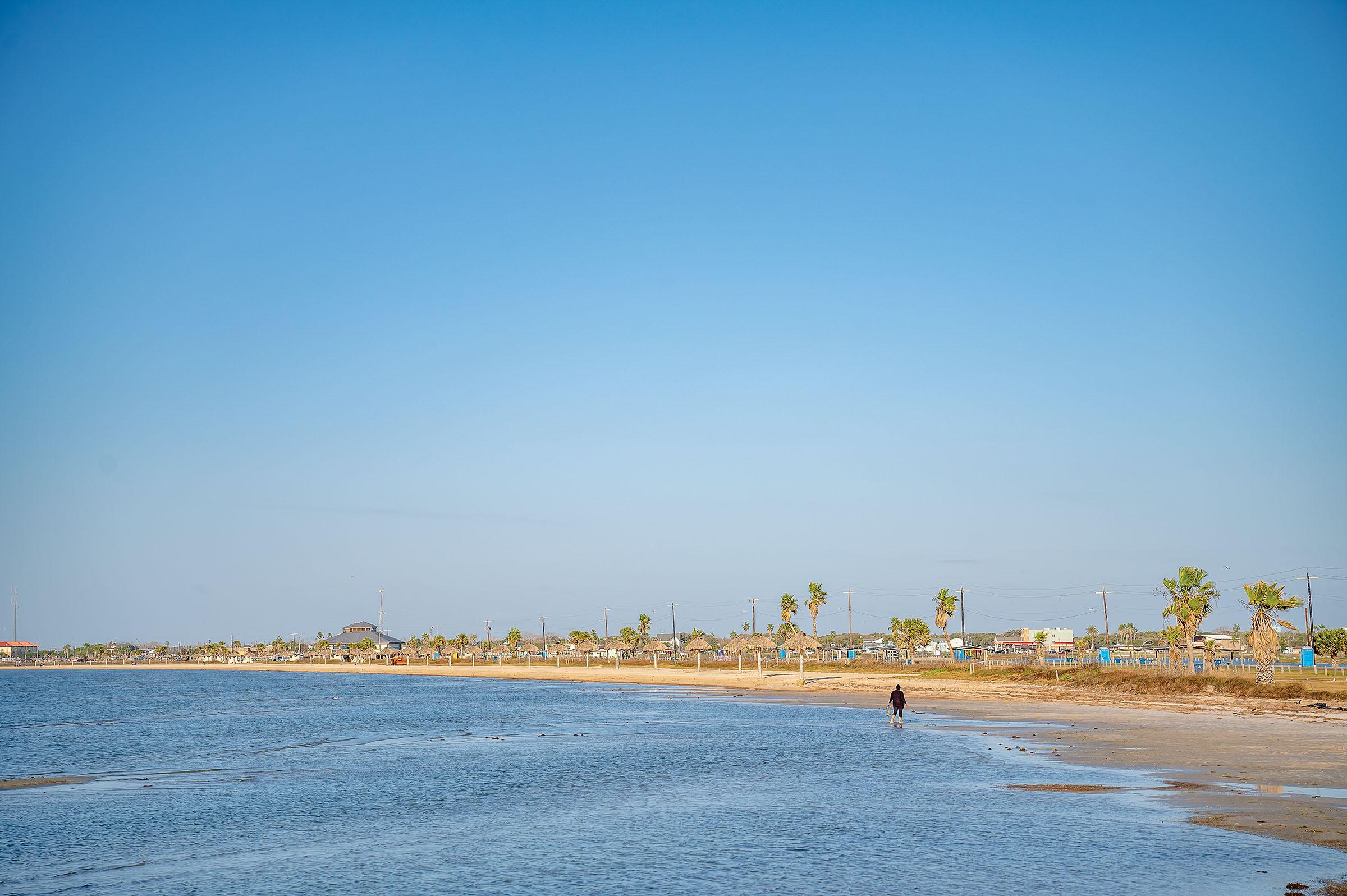 A person walks the sandy shore next to a large body of water near palm trees and wooden shade structures