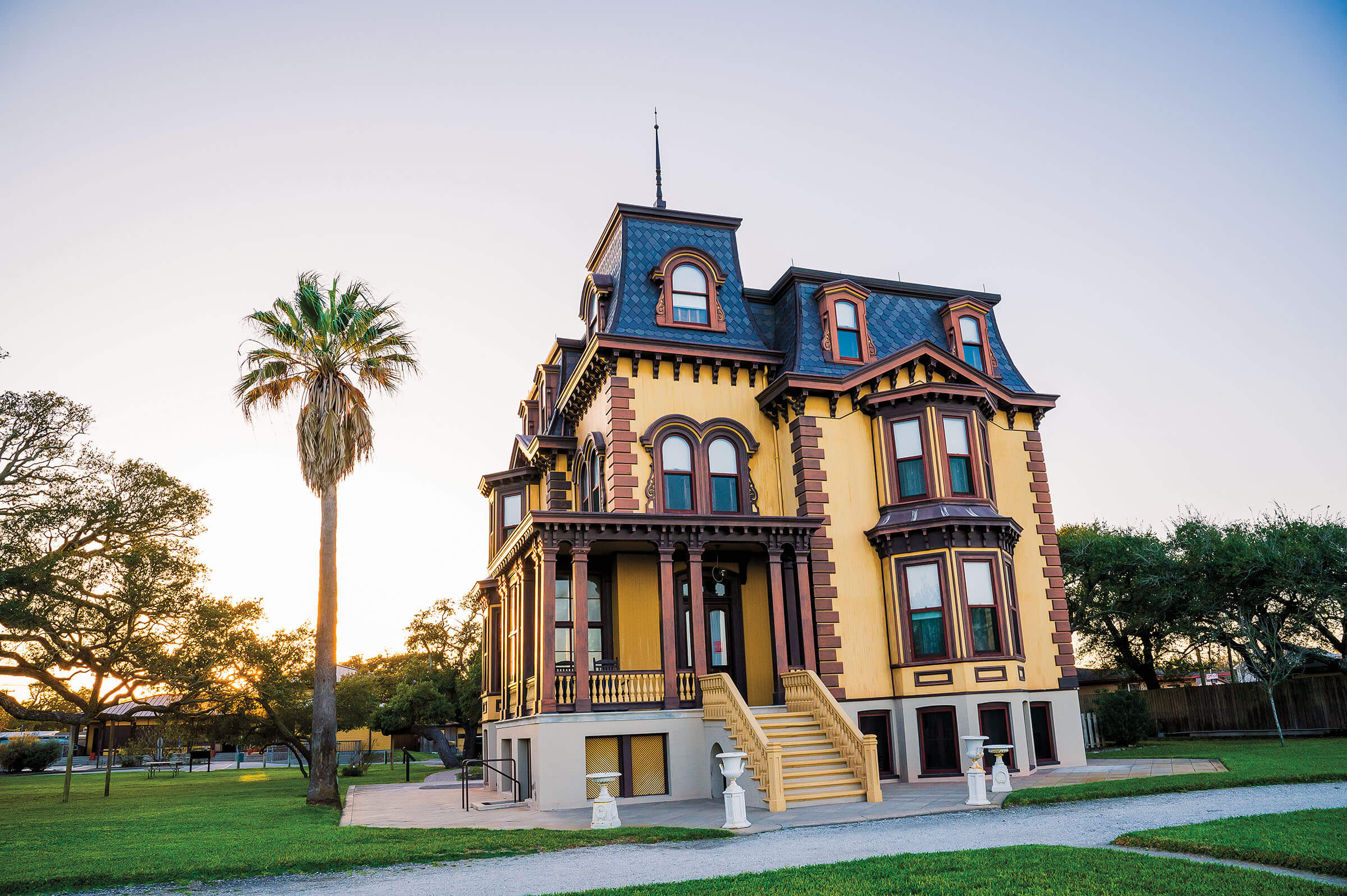 A large yellow Victorian-style home with a dark roof next to a tall palm tree