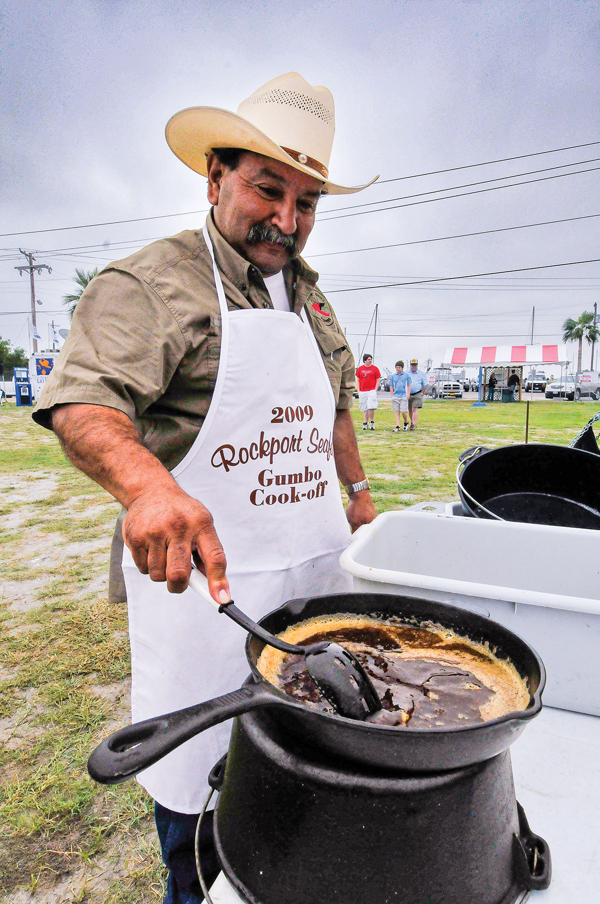 A man in a cowboy hat and white apron lowers food into a large cast-iron pan