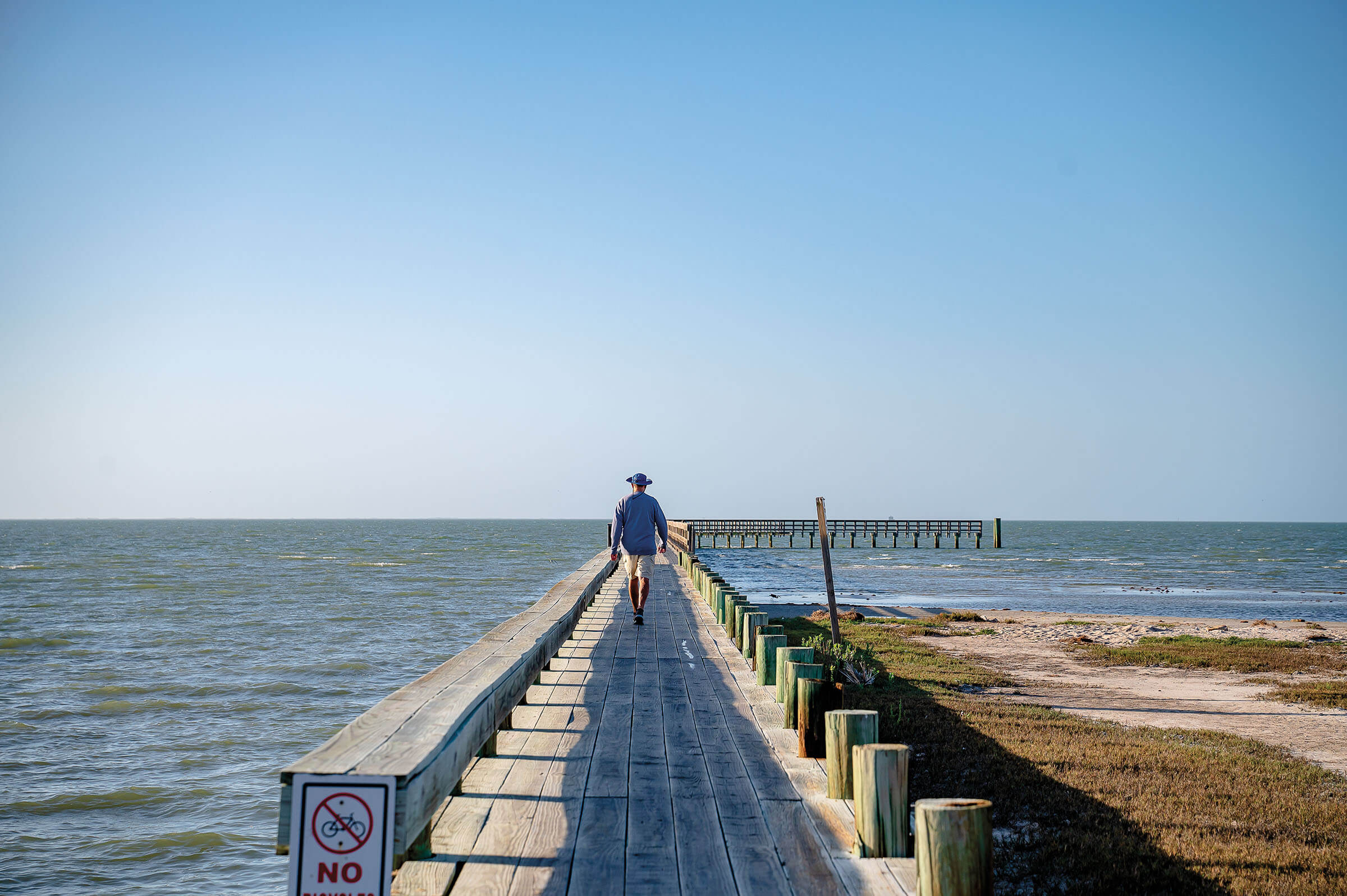 A person walks down a long wooden pier above blue water and under blue sky