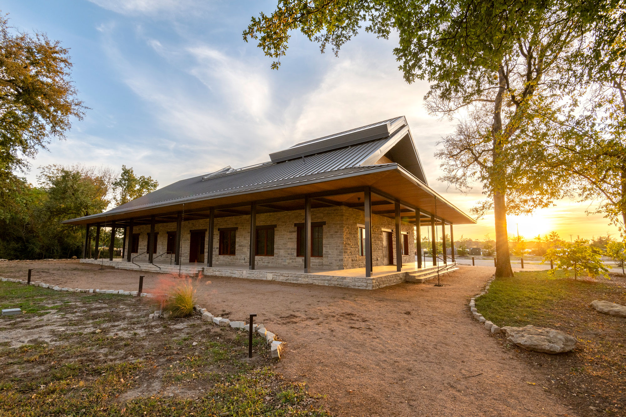 A building with a metal roof and limestone bricks stands on a plot of land with a gravel track leading to it.