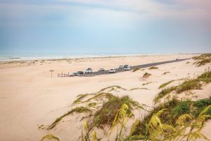 Cars park on a road that ends and leads into sand dunes with an ocean in the background