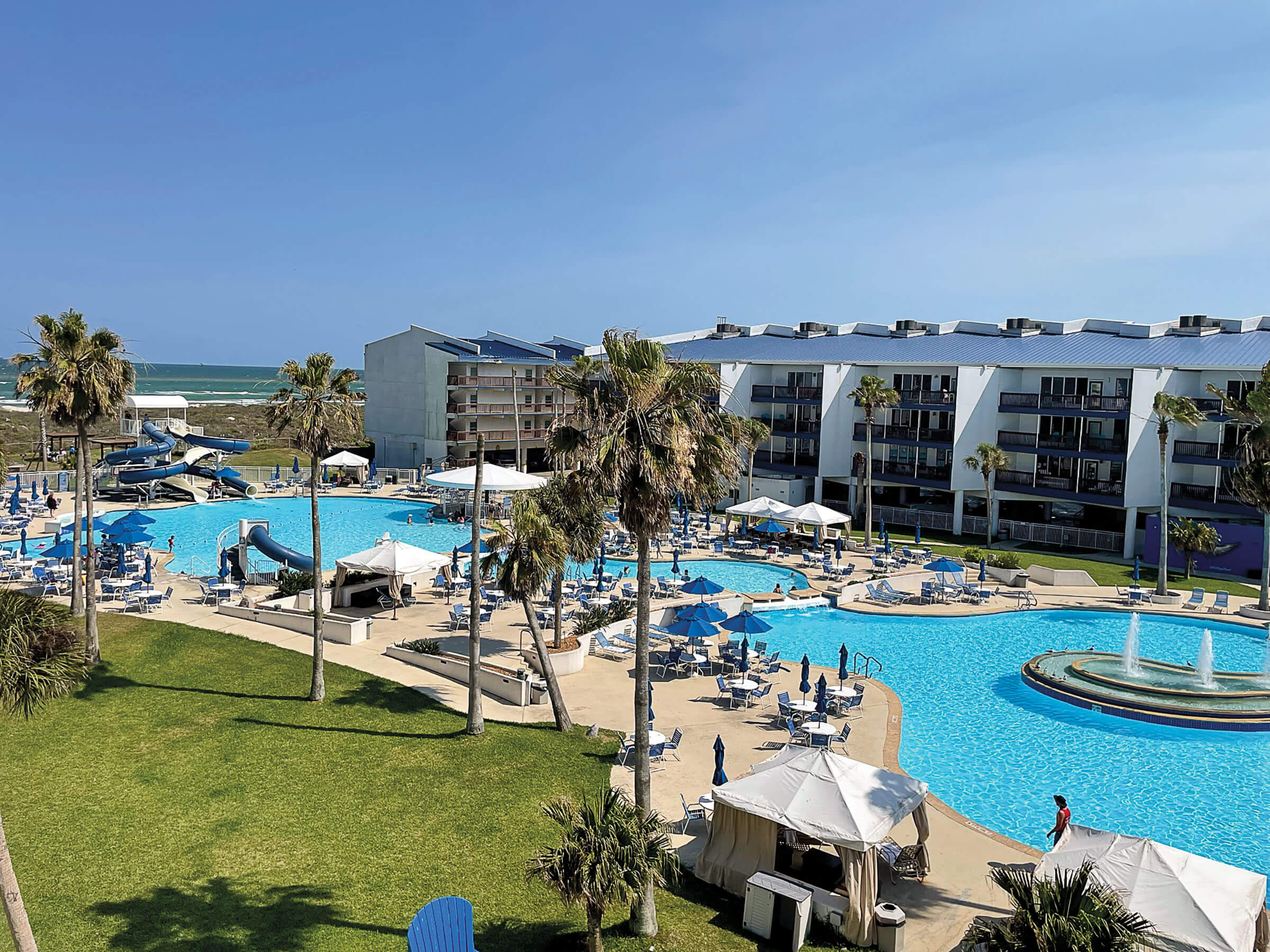 An overhead view of a resort with low-rise hotel room buildings and a very large, bright blue pool and palm trees