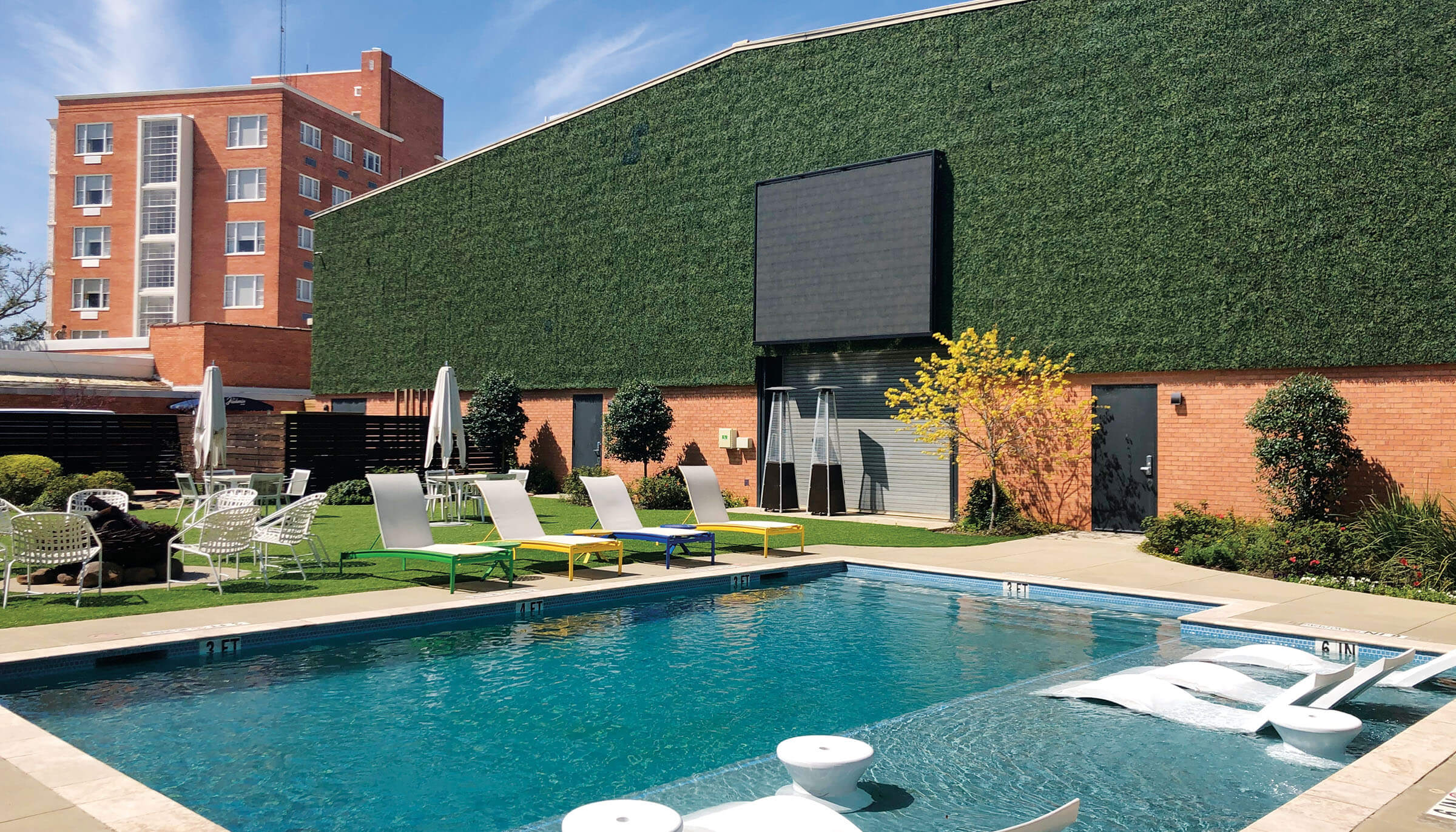 A TV screen mounted against a green backdrop over a large blue pool with lounge chairs